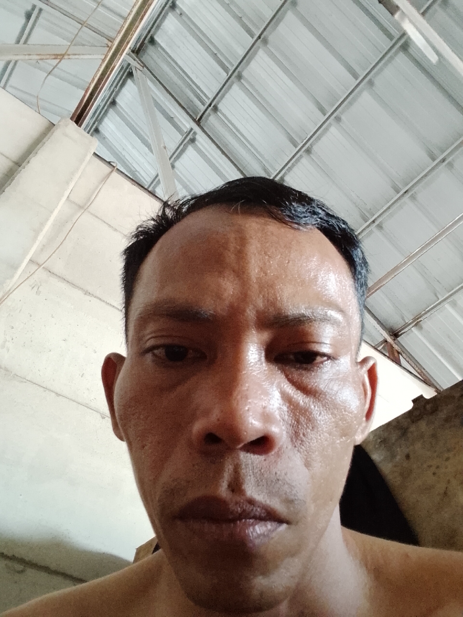 The image is a close-up, low-angle selfie of an adult male. The man's face is the central focus, with his forehead, eyes, nose, and mouth visible. His skin appears to be sweaty or oily, with visible pores and some texture. He has short, dark hair. His gaze is directed downwards, away from the camera.
The background consists of an indoor environment with a corrugated metal ceiling and exposed metal beams. The walls appear to be a light color, possibly concrete or plaster. The lighting suggests an indoor space, possibly with natural light entering from above or from the side.
The image was taken in Bukit Ketok, Indonesia.