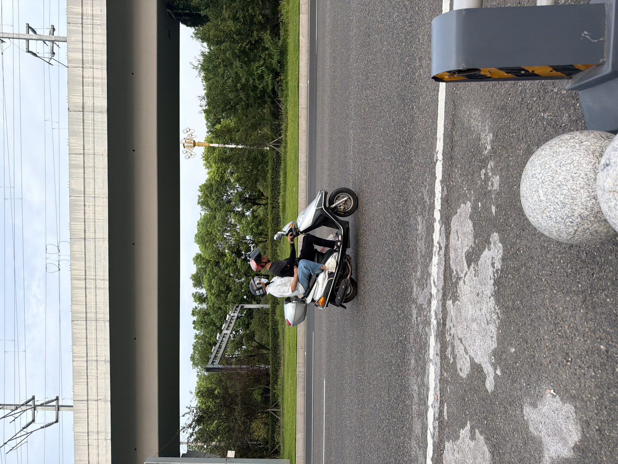 Two individuals wearing helmets ride on a scooter on a paved road in Jiangjin, China. The scooter is silver and black with a storage box on the back. The individuals are positioned one behind the other, with the person in front wearing a white t-shirt and blue jeans, and the person in the back wearing a dark t-shirt and dark pants. They are both looking forward. The road has a white lane marking. In the background, there is a grassy area with trees and a raised concrete structure overhead with power lines. A lamppost with multiple lights is visible among the trees. The sky is overcast. In the foreground, there are two spherical objects and a gray structure with yellow and black stripes, suggesting a traffic barrier or bollard.