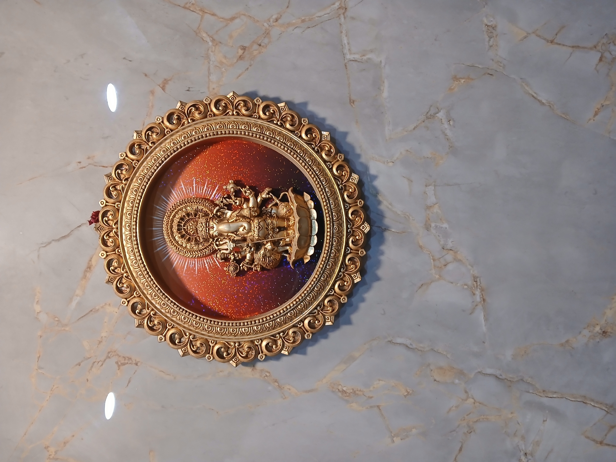The image shows a circular, ornate gold-colored frame mounted on a gray and white marble-patterned wall. Inside the frame is a reddish-orange background with glitter-like sparkles. A golden deity statue, possibly Vishnu or Lakshmi, is positioned within the frame. A reflection of a light source is visible on the wall above the frame. The precise location in Bengaluru, India cannot be determined from the image.