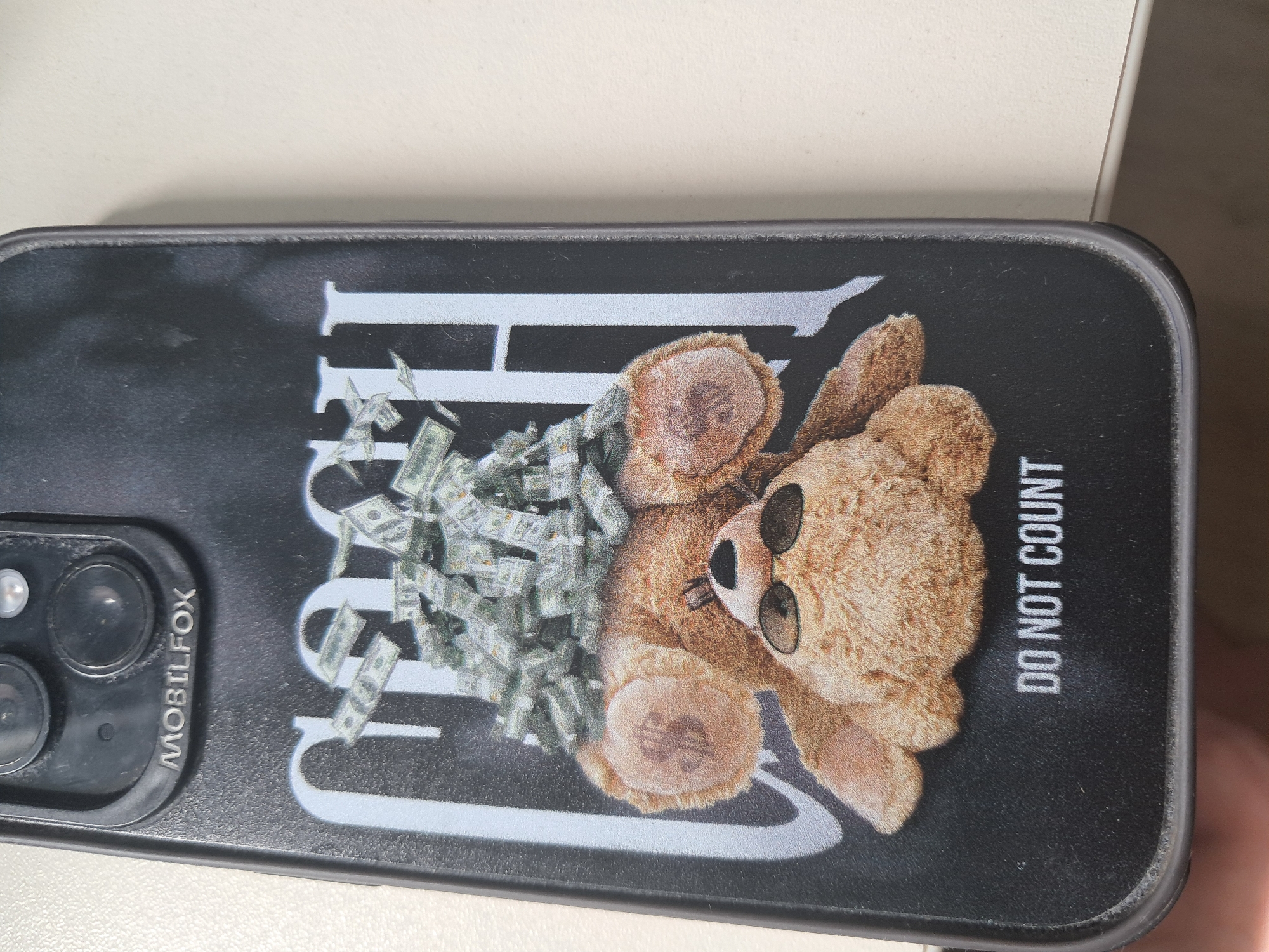A black smartphone case, bearing the "MOBILFOX" brand name near the camera module, is depicted. The case features a graphic design: a light brown teddy bear wearing dark sunglasses is lying on its back. A substantial pile of green currency notes, resembling US dollars, covers the teddy bear's torso and lower head. Above the bear, partially obscured by the currency, are the large, light grey letters spelling "CASH". Below the bear, in smaller light grey lettering, the phrase "DO NOT COUNT" is visible. The phone's camera housing is dark, containing three distinct lenses and a flash. The object is held by an individual, with fingers visible at the bottom-left corner, and rests on a light, uniform surface. The scene does not contain visual information to determine a specific location within the city of Plovdiv, Bulgaria.