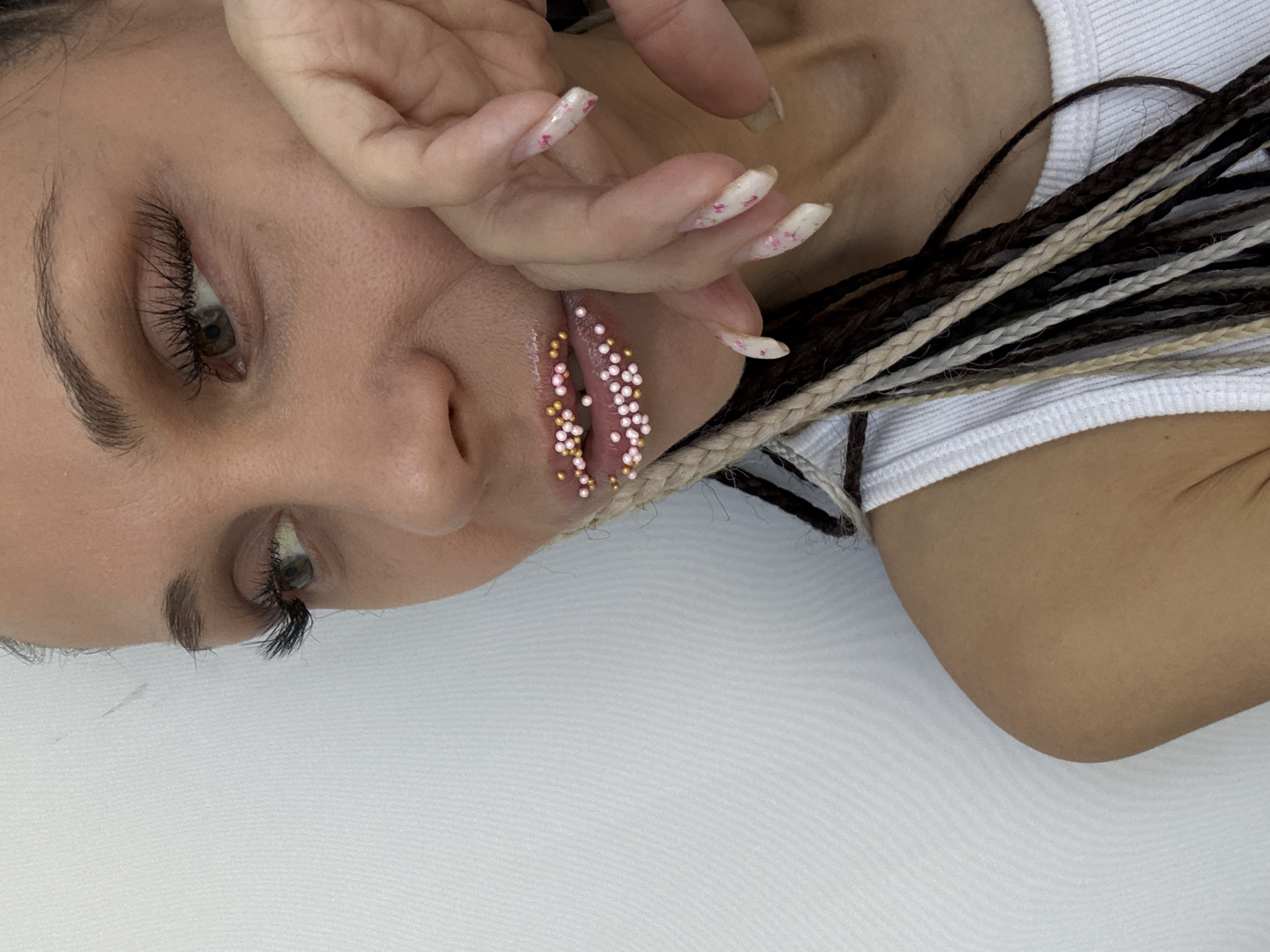 A close-up image depicts a person with brown and blonde braided hair. The individual is wearing a white shirt. Their lips are adorned with small, round, pink and gold decorations. Their hand, with long nails painted with a white and pink design, is near their face. The background is a plain, white surface. The location is unknown.