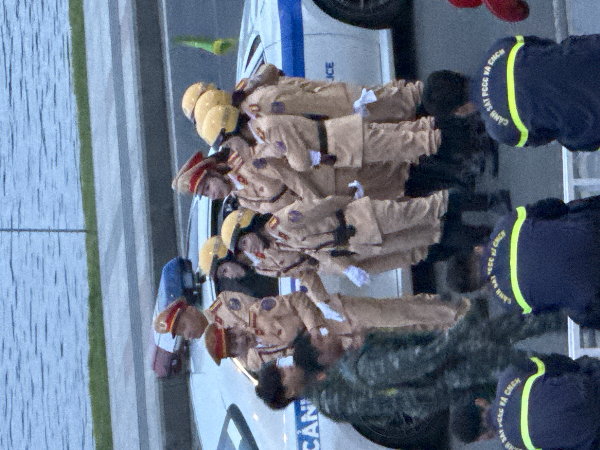 In this medium-distance, eye-level shot, a group of individuals in uniform are gathered near a white car with a blue and white stripe. The car has "CAN" visible on its side. The individuals are wearing tan uniforms with red caps. Some of them are also wearing yellow helmets. There are at least seven people in uniform visible in the foreground and midground. Two individuals in dark blue uniforms with yellow reflective stripes are seated in the lower foreground, facing away from the camera, with text on their backs. The environment appears to be outdoors, with a body of water and a grassy area in the background, suggesting a location near Xuan Huong Lake in Da Lat, Vietnam. The lighting suggests it is daytime. The individuals appear to be part of a formal event or ceremony.
