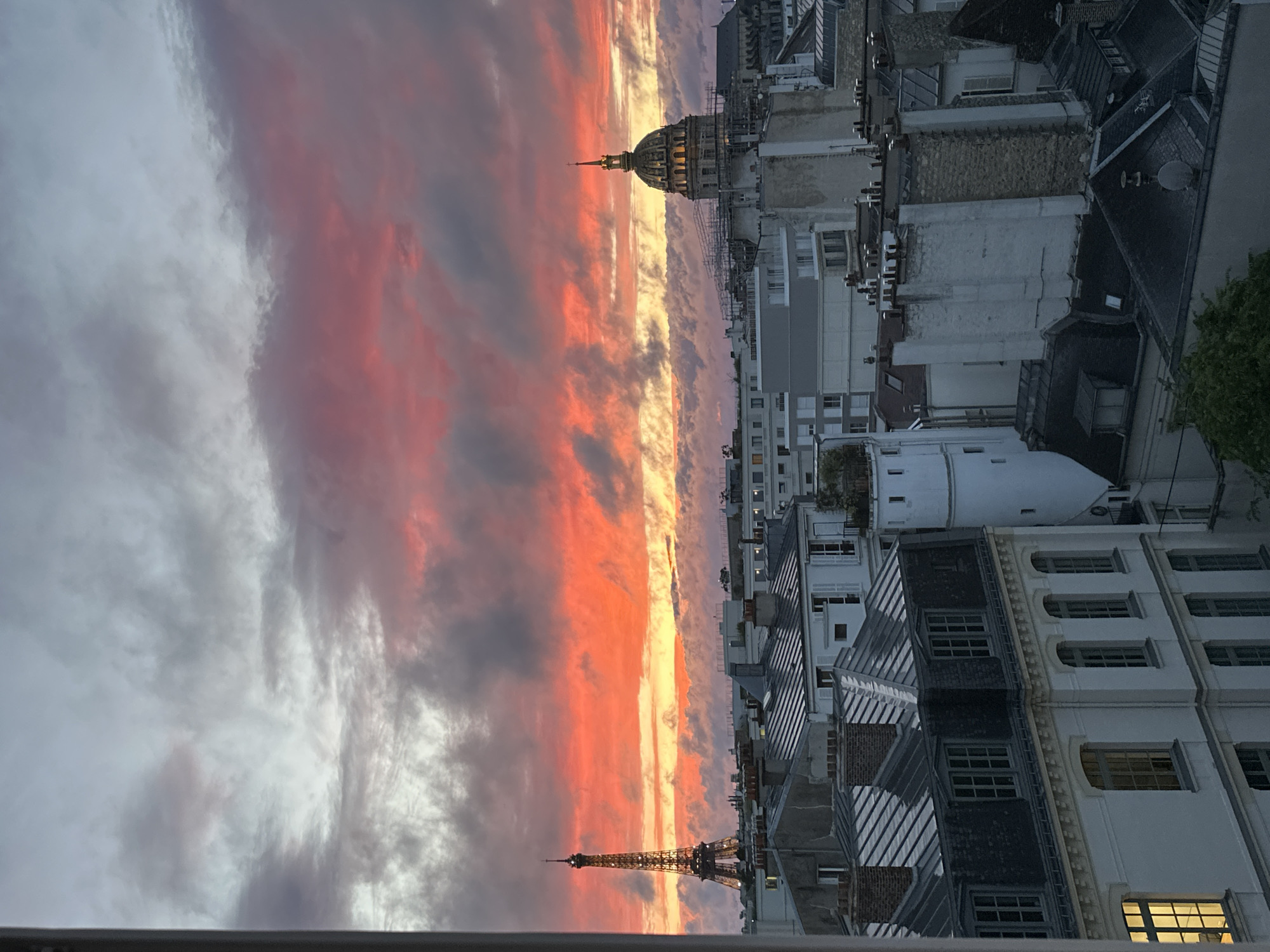 The image presents a breathtaking rooftop view of Paris at sunset, oriented as if looking towards the west. The sky is dominated by a dramatic display of colors, with vibrant streaks of fiery red, orange, and yellow clouds stretching horizontally across the horizon, contrasting sharply with cooler grey and light purple clouds above.
On the left side of the frame, the iconic Eiffel Tower stands tall, its recognizable silhouette piercing the colorful sky. Towards the center-right, the distinct dome of a significant Parisian structure, likely Les Invalides or the Pantheon given the context and architectural style, is visible, also silhouetted against the bright hues of the sunset. Some scaffolding can be observed around the dome.
The lower half of the image is filled with a dense array of traditional Parisian buildings, characterized by their mansard roofs, chimneys, and dormer windows. These buildings are largely cast in shadow due to the twilight, accentuating the vibrant illumination of the sky. There is no specific activity taking place, and no discernible text is visible within the scene. The overall impression is one of serene beauty, capturing the transition from day to night over the historic cityscape.