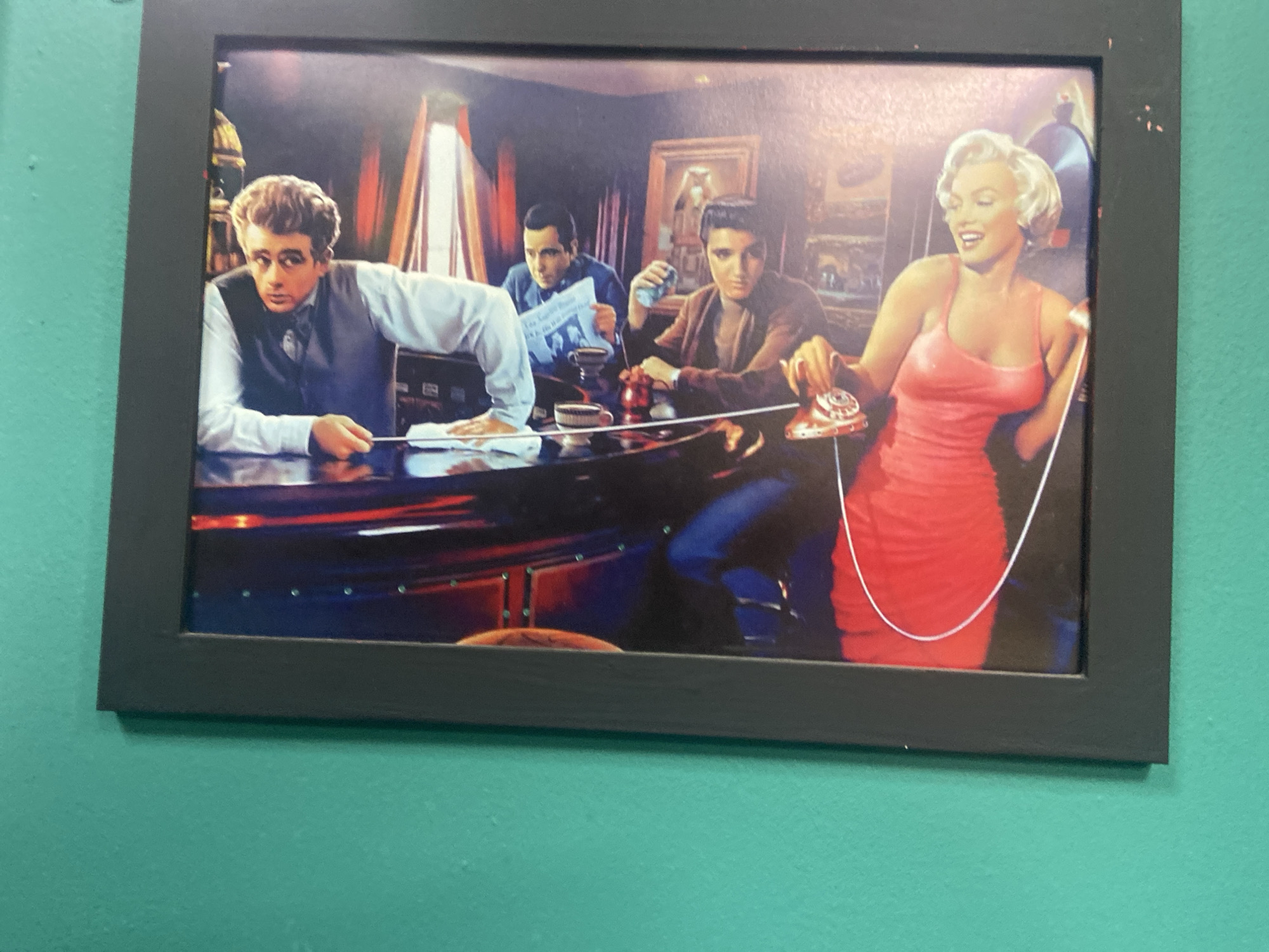 This is a framed photograph depicting four individuals in a dimly lit, possibly retro-themed interior. The photograph appears to be a composite image or an artistic rendering, not a candid snapshot.
On the left, a man resembling James Dean is seated at a dark, polished counter, looking intently to his right. He is wearing a white dress shirt, a dark vest, and a dark tie. His left hand is resting on the counter, holding a small object, while his right arm is bent, and his hand is also on the counter.
To his right, another man, possibly resembling Marlon Brando, is seated, holding a newspaper and a cup. He is looking down and appears to be reading.
Further to the right, a man who resembles Elvis Presley is seated, holding a light blue cloth in his left hand and reaching with his right hand towards a red rotary telephone. He is looking towards the right side of the frame.
On the far right, a woman who strongly resembles Marilyn Monroe is seated, wearing a bright pink, form-fitting dress. She is smiling and looking towards the left, with her right hand holding the receiver of the red rotary telephone. A white cord from the telephone extends across the image.
The background features what appears to be a bar or lounge setting, with dimly lit walls, framed artwork, and curtains. The lighting is dramatic, casting shadows and highlighting the figures.
The photograph is framed in a black, rectangular frame. The image is mounted on a teal-colored wall.
The prompt mentions the city of Yegorlykskaya, Russia. However, there are no visual cues within the image itself that indicate this specific location. The scene depicted is generic in its setting and does not contain any identifiable landmarks, signage, or cultural elements that would place it in Yegorlykskaya. Therefore, this location information is not supported by the visual content of the image.