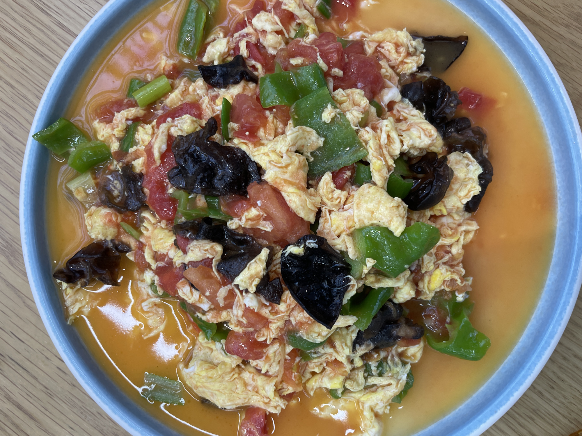 A top-down, medium shot captures a plate of Chinese tomato and egg stir-fry, garnished with wood ear mushrooms and green peppers. The dish sits in a light-orange sauce, presented on a blue-rimmed plate. The scene appears to be indoors, with a light-colored wooden surface visible underneath the plate. There is no indication of the location being in Lintong District, China. The image focuses solely on the food and its immediate surroundings.