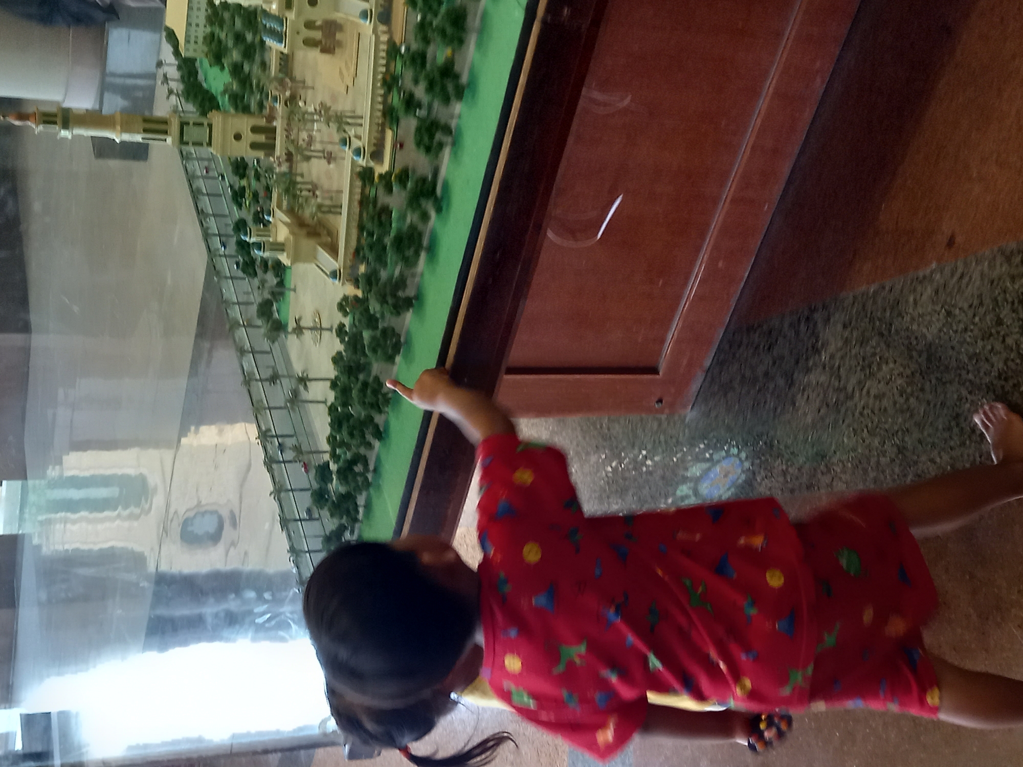 The image captures two young children, a boy and a girl, interacting with a miniature model of a mosque. The boy, wearing a red shirt with a dinosaur print and red shorts, is positioned in the foreground, his right arm extended as he points at the model. He is barefoot. The girl, with her hair in a ponytail and wearing a yellow shirt, stands to his left, partially visible. They are inside a building with a polished floor that reflects the surrounding environment, suggesting an indoor exhibition or museum setting. The mosque model features intricate architectural details, including minarets, domes, and surrounding greenery, resembling a scaled-down representation of a significant religious structure. The overall scene is brightly lit, with a focus on the children and their engagement with the exhibit. This scene is located at the Islamic Center Samarinda in Samarinda, Indonesia.