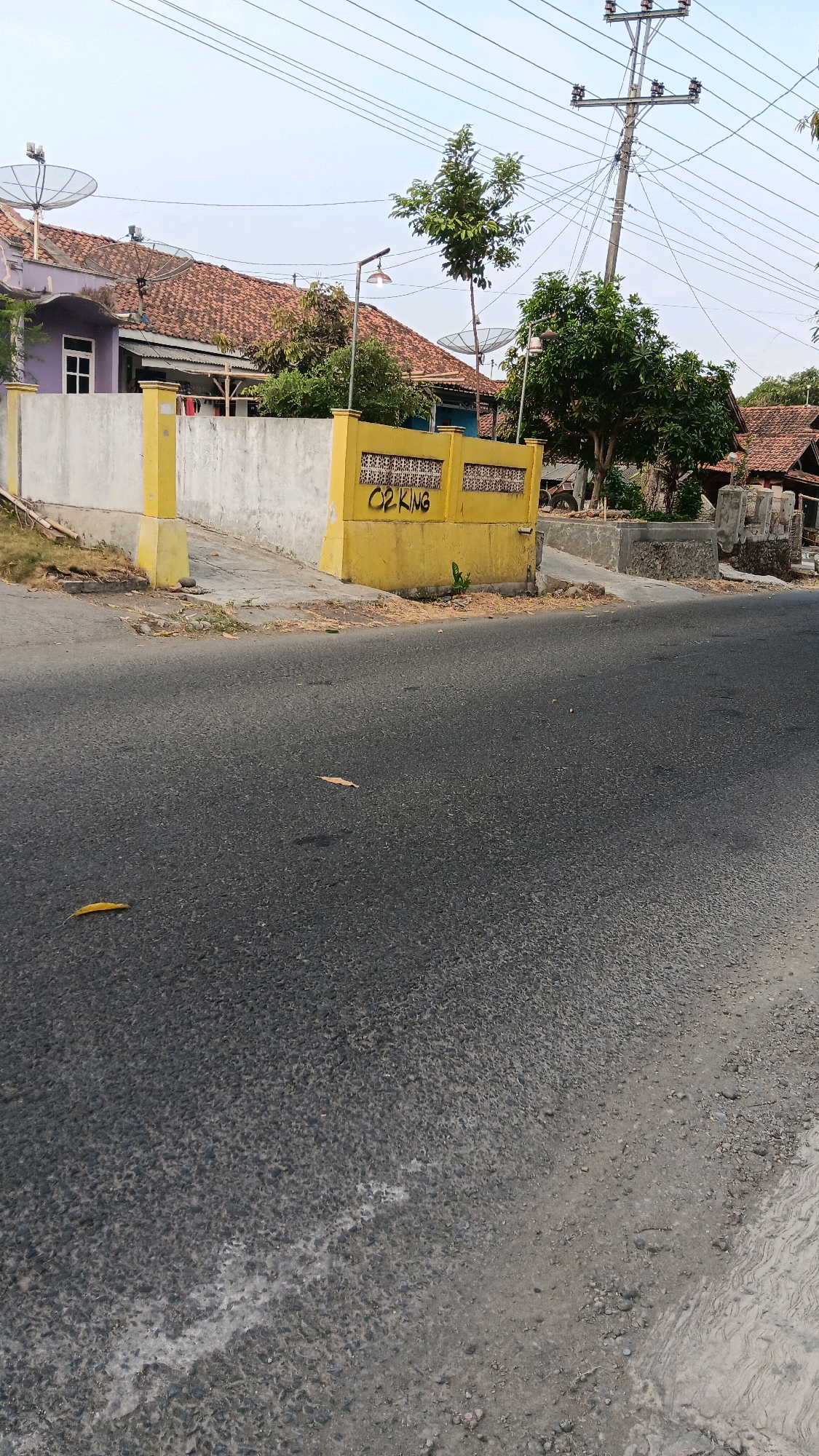 In Pangebatan, Indonesia, a roadside scene unfolds with a paved asphalt road in the foreground, showing signs of wear and scattered dry leaves. Adjacent to the road is a concrete pathway leading to a yellow painted wall and structure. The yellow structure, adorned with patterned openings, bears graffiti in black marker that reads "O2 KING". Behind the yellow structure, a white concrete wall extends to the left. Further in the background, a purple-colored house with a red tiled roof is visible, featuring a white-framed window. Several satellite dishes are mounted on the roofs of the houses and on poles. Trees of varying sizes dot the landscape, and a utility pole with numerous wires stands prominently against the sky. The overall environment appears to be a residential area bordering a road, possibly in a rural or semi-urban setting.