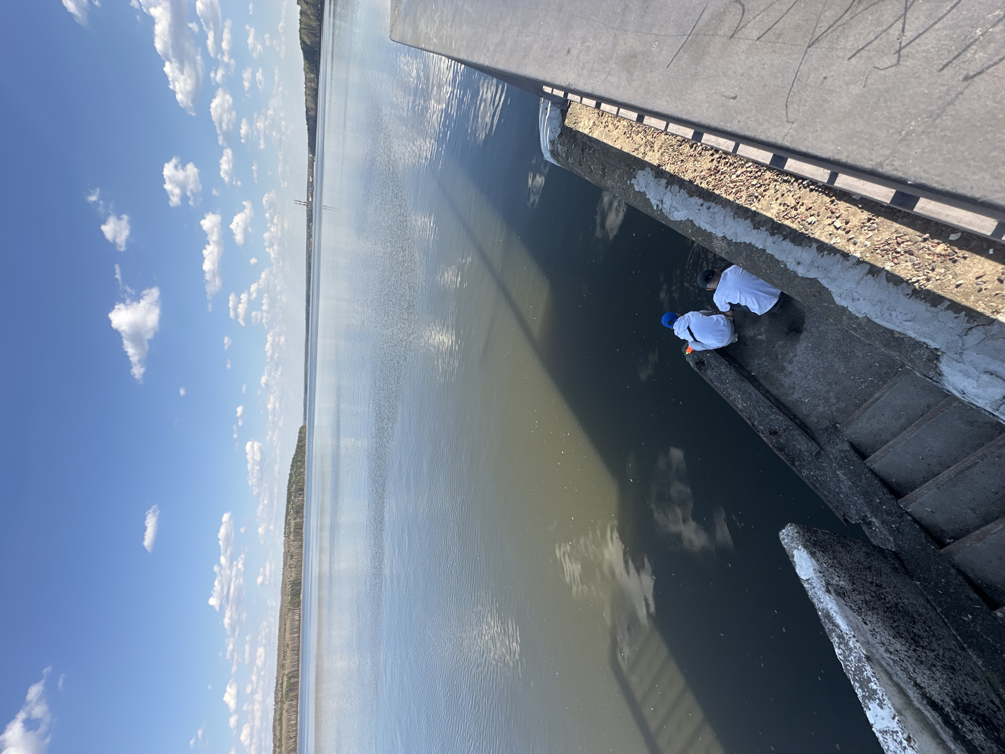 The image shows an elevated view of a waterway with a concrete structure. Two people are located on a concrete ledge near the water's edge, both wearing white shirts. One individual also has a blue cap. The water reflects the sky and clouds above. A narrow strip of land is visible in the distance. The location is identified as Chaikovsky, Russia, though specific landmarks are not discernible.