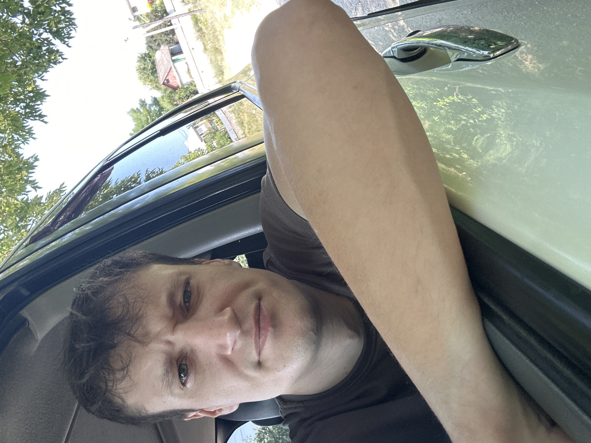 The image shows a man leaning out of a car window, with his right arm resting on the car door. He is looking towards the camera with a neutral expression. The background shows a rural scene with houses, trees, and a bright sky. The car appears to be parked on a dirt road. The man is wearing a dark brown t-shirt. The location is identified as Makiv, Ukraine.