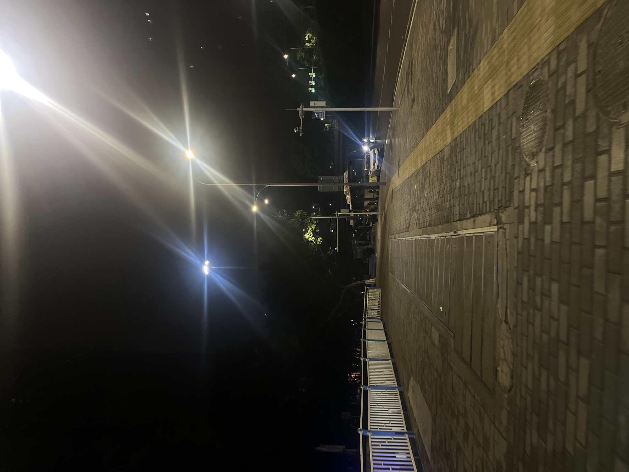 This image captures a nighttime urban street scene in Jiulongpo District, China. The perspective is from a low vantage point on a paved sidewalk, looking forward alongside a road.
On the left side of the image, a white and blue metal railing extends into the mid-ground, bordering a dark, vegetated area. The sidewalk itself is composed of dark grey interlocking pavers, with sections of worn, lighter-toned pavement and embedded linear metal strips.
On the right, the sidewalk continues with similar grey pavers, featuring a continuous yellow tactile paving strip running parallel to the road, extending into the mid-ground. Several circular metal manhole covers are visible on the right sidewalk.
A multi-lane asphalt road runs parallel to the sidewalk on the right. In the mid-ground, a dark-colored bus is stationary on the road's left side. Behind the bus, a white sedan is also stopped, with its headlights brightly illuminated, projecting light forward. Near the white sedan, an indistinct figure of at least one person is visible, illuminated by the car's headlights.
Multiple streetlights illuminate the scene, creating significant lens flares and light streaks, characteristic of nighttime photography. A tall street light pole with a bright white light stands on the sidewalk between the bus and the white car. Further down the road, another pole supports a surveillance camera and a sign with a camera icon. Green traffic lights are visible overhead in the distance. The areas beyond the road and the railing are dark, revealing the silhouettes of trees and distant, faint artificial lights suggesting urban development under a dark night sky.