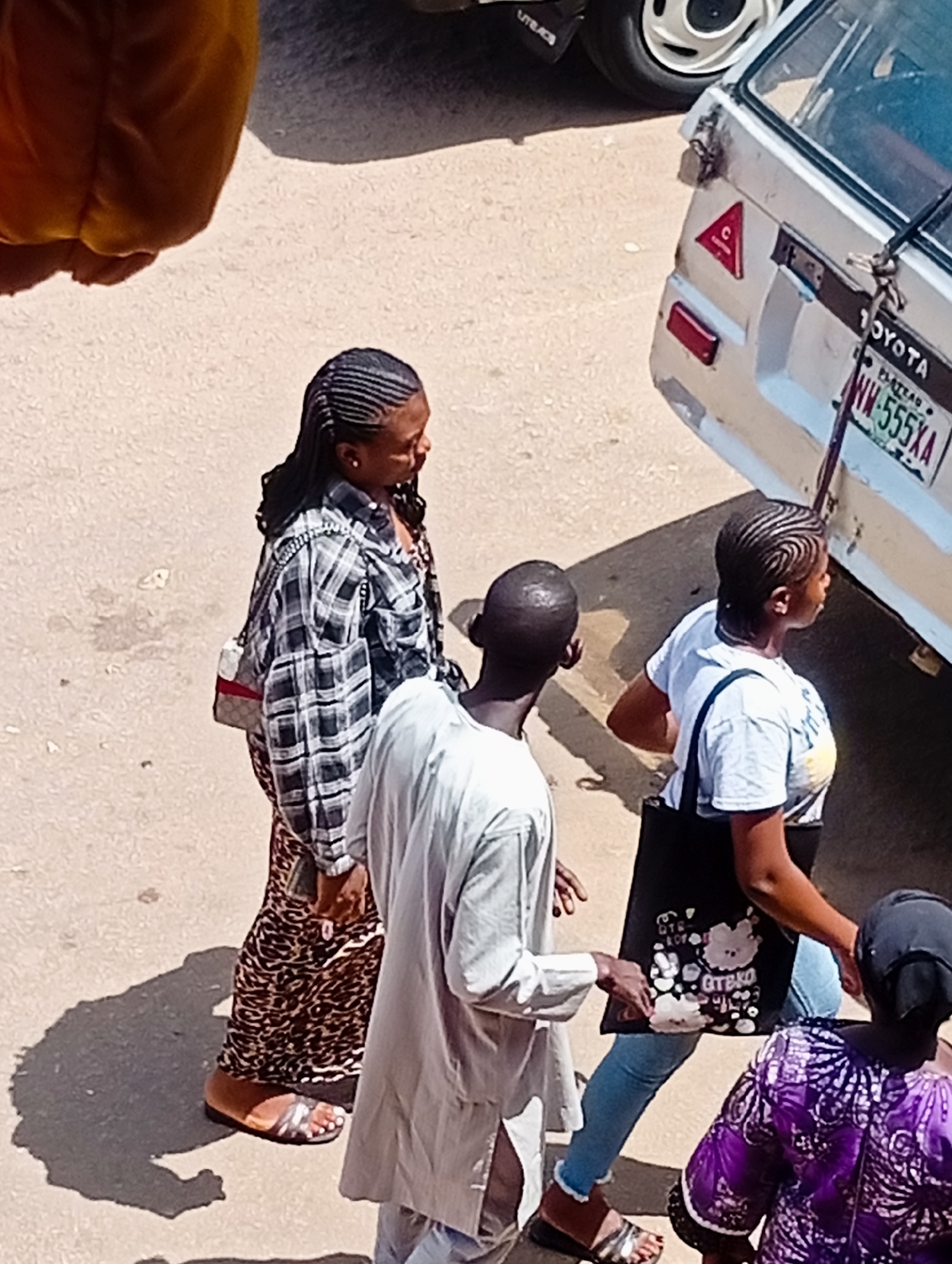 The image captures a street scene in Bukuru, Nigeria, with several individuals and a vehicle. In the foreground, three people are visible, two women and a man. One woman, on the left, wears a plaid shirt and a patterned skirt, carrying a small handbag. She has her hair styled in thin braids. A man in a light-colored tunic stands beside her. To the right, another woman, also with braided hair, is wearing a light t-shirt and blue jeans, carrying a black tote bag. Further in the background, the rear of a white Toyota vehicle is prominent, showing a license plate with the text "NW-555XA" and a triangular red warning sticker. A person in a purple patterned garment is partially visible in the lower right corner. The scene appears to be outdoors, with sunlight casting shadows, suggesting it's daytime. The ground is paved and appears dusty. The overall impression is that of people on a street, possibly walking or waiting near a vehicle.