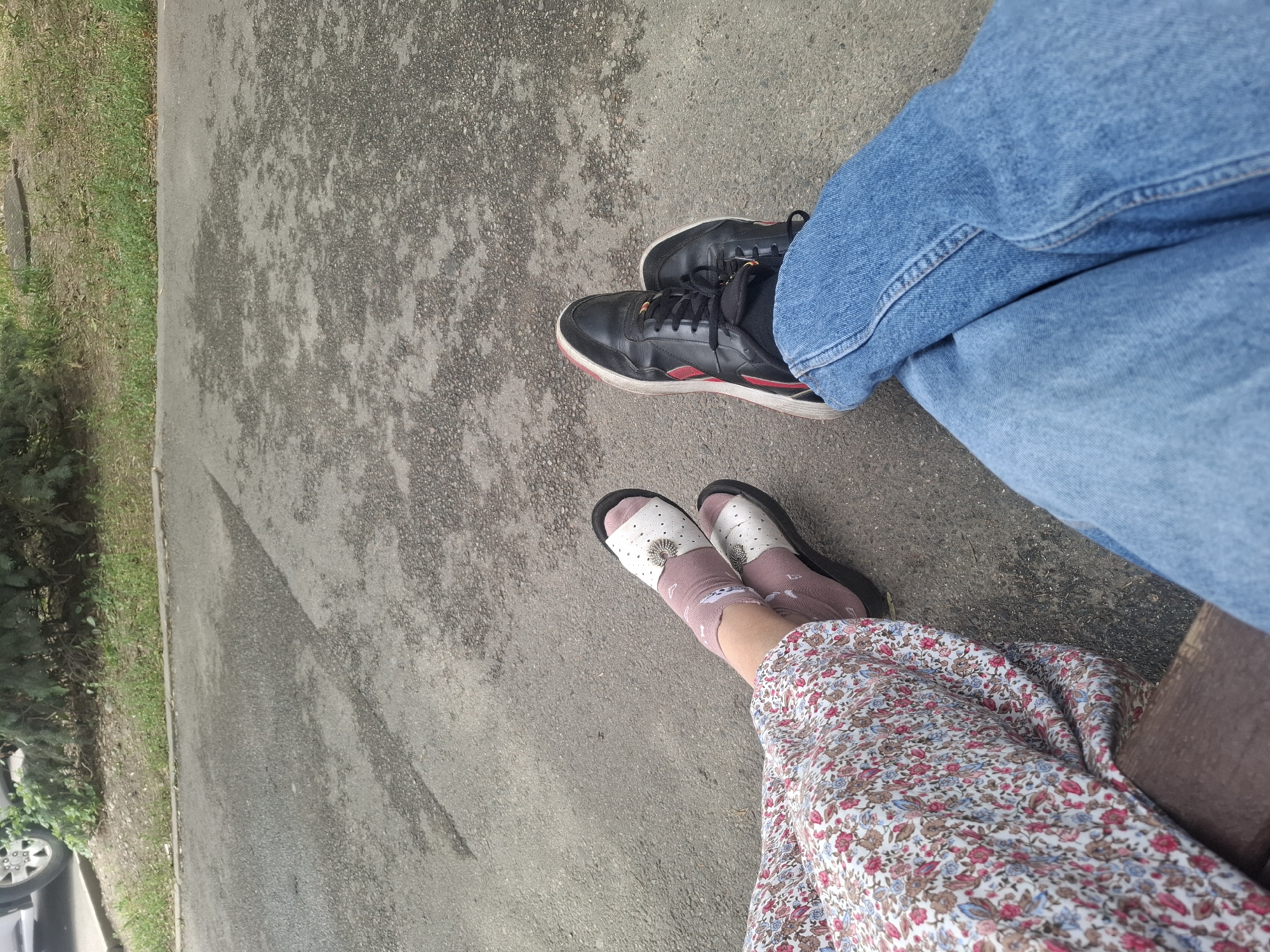 Two individuals are seated on a bench outdoors. The person on the left is wearing a floral-patterned dress and white sandals with decorative buckles, paired with pink socks. The person on the right is wearing blue jeans and black athletic shoes with red accents. They are both sitting with their legs crossed. The ground is paved with asphalt. In the background, there is a patch of grass and trees, suggesting a park or a green space within the city. The image does not contain enough information to identify the specific location in Almaty, Kazakhstan.