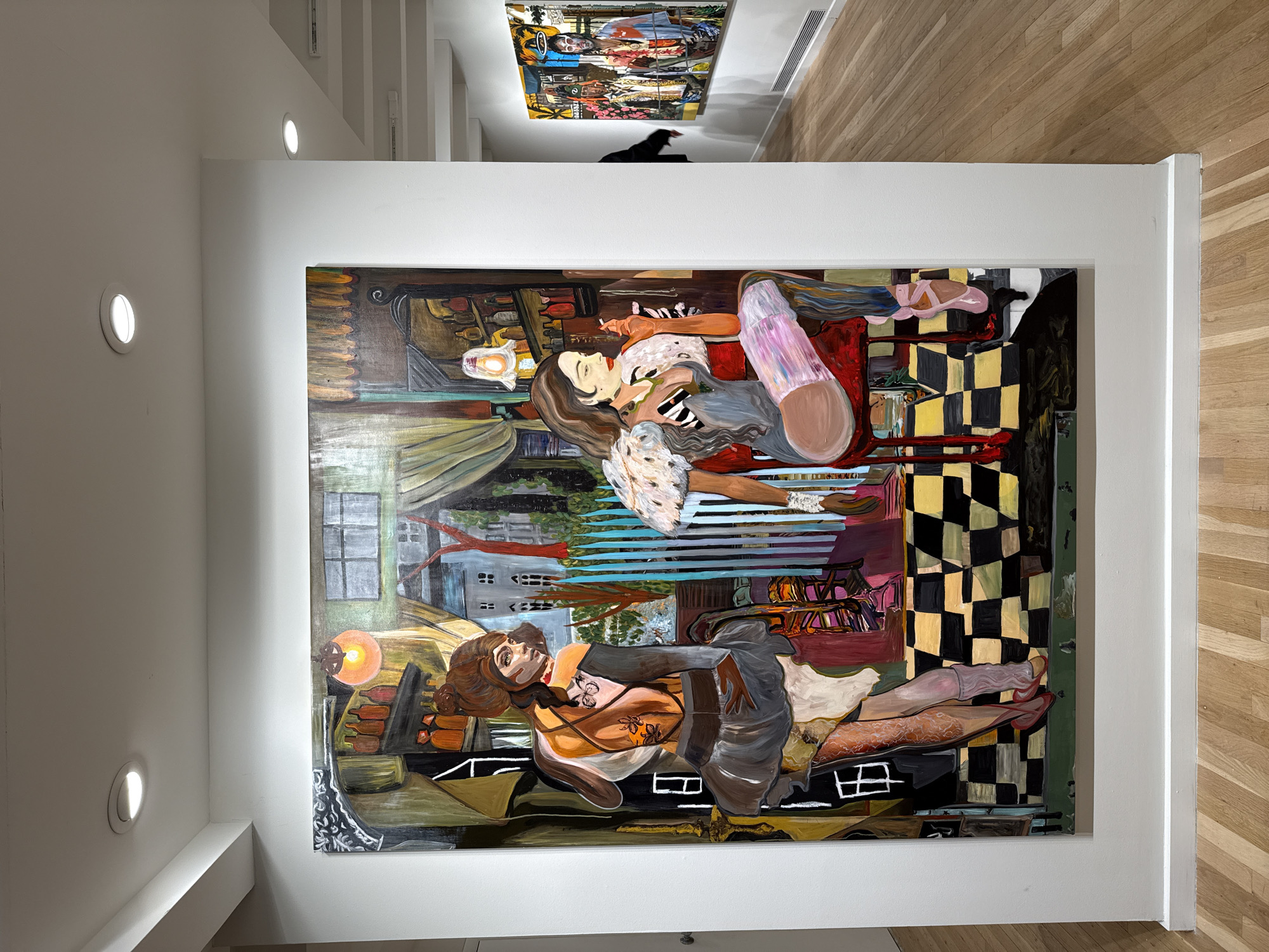 The image is a photograph of a painting displayed in what appears to be an art gallery, potentially located in New York City based on the setting. The painting features two women lying in different positions, depicted in a slightly surreal or dreamlike style. The background is a mix of interior and exterior elements, including furniture, checkered patterns, and what might be windows with a view of buildings or trees. The painting itself is framed and hung on a white wall in a well-lit space with recessed lighting in the ceiling. The floor is made of wood. Another painting is visible on a distant wall, partially obscured by the ceiling's structure. A figure wearing black is slightly visible beneath this painting, standing in the background.
