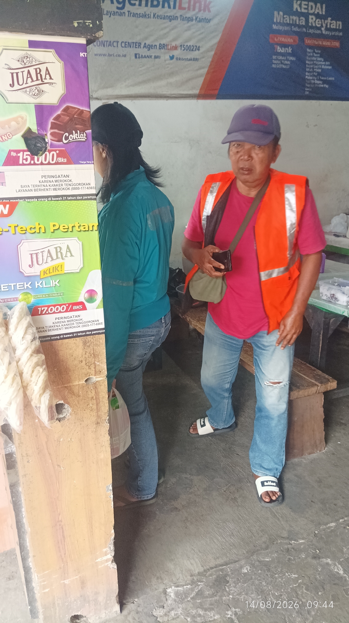 The image shows an outdoor scene with two individuals. On the left, a person wearing a teal jacket and jeans is standing, partially obscured by a wooden post. They are looking to the right, towards the second individual. On the right, a person wearing a pink t-shirt, blue jeans with a ripped knee, and an orange safety vest over it is standing and facing forward. This person is holding a small dark wallet or cardholder in their right hand and has a crossbody bag slung over their shoulder. They are wearing white slide sandals. The background shows a banner with text in Indonesian, including "AGEN BRILINK," "LAYANAN TRANSAKSI KEUANGAN TANPA KANTOR," and "KEDAI Mama Reyfan." Another banner to the left has the word "JUARA" prominently displayed, along with images of chocolate and crackers, and pricing information like "Rp. 15.000/BKS." The ground is concrete. A wooden bench is visible behind the person in the orange vest. The overall setting appears to be an outdoor market or a small shop in Tasikmalaya, Indonesia, given the mention of "AGEN BRILINK" and "KEDAI Mama Reyfan," which are common services and businesses in Indonesian cities. The date and time stamp on the image indicates "14/08/2026 09:44."