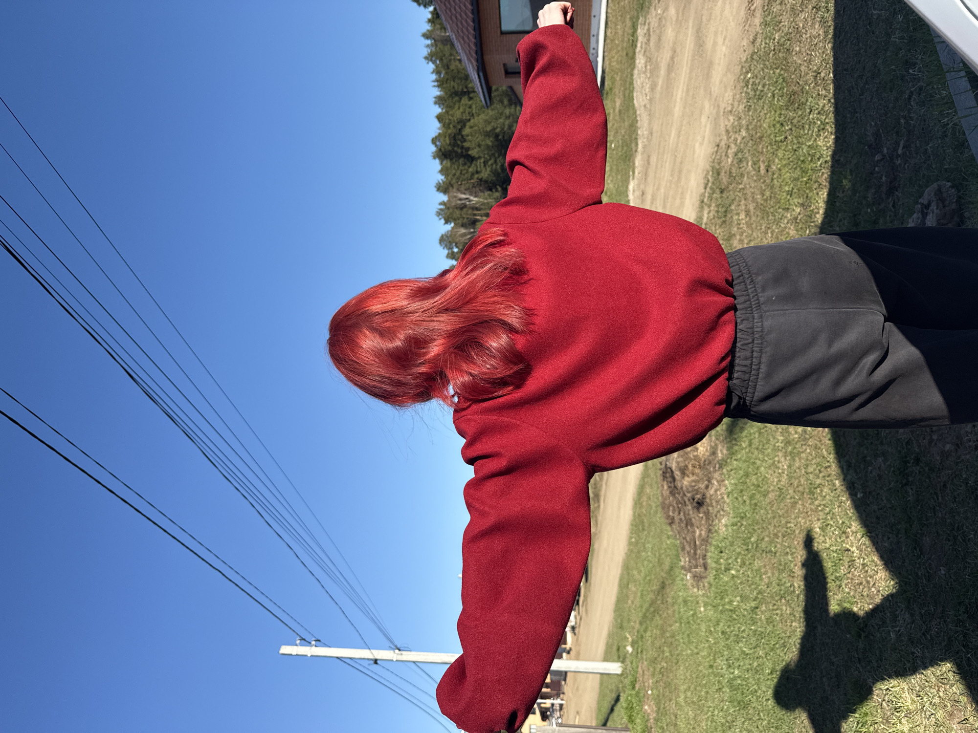 A person with red hair is seen from the back, wearing a red jacket and black pants. They are standing outdoors on a sunny day with a blue sky. Power lines are visible in the upper left. There is grass and a dirt path in the background, as well as some trees and a building. The location is Chaikovsky, Russia.