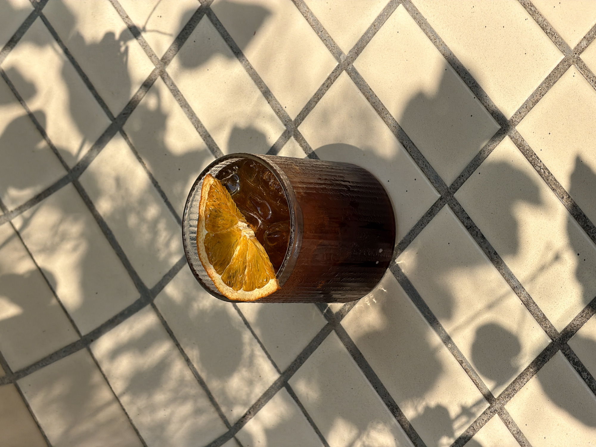 A high-angle shot shows a clear, textured glass of dark liquid with ice and an orange slice, resting on a light-colored tiled surface with dark grout lines. Shadows of leaves or foliage are cast across the surface.
The specific location in Đà Nẵng, Vietnam, cannot be determined from this image.