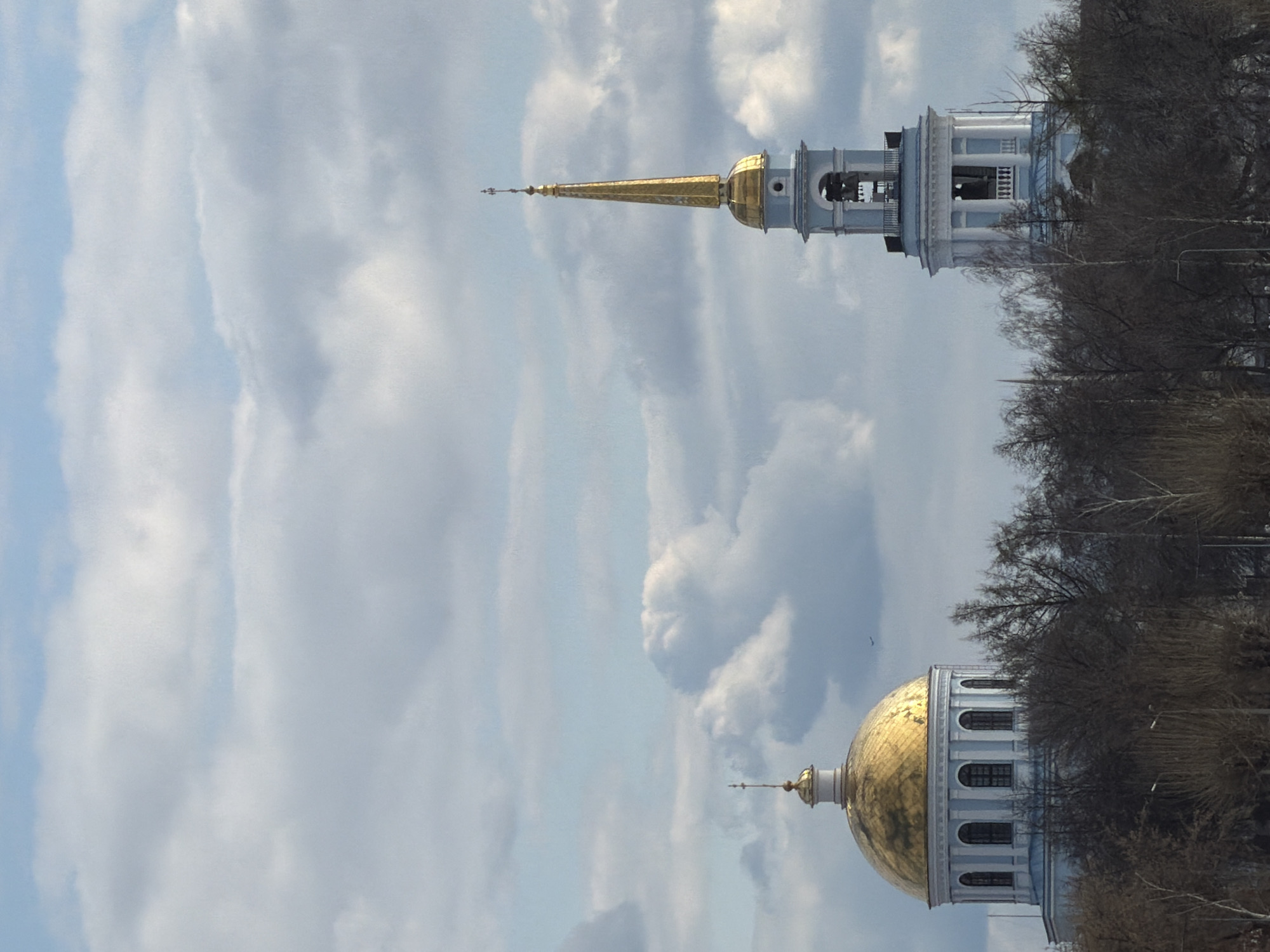 The image, appearing to be rotated 90 degrees counter-clockwise, depicts two distinct architectural structures, likely part of a church or cathedral complex, set against a cloudy daytime sky. When viewed in its intended upright orientation, the structures are on the right side of the frame, partially obscured by bare trees.
The upper structure features a prominent, slender golden spire topped with an ornate finial. Below the spire is a smaller golden dome, leading down to a white bell tower with arched openings housing bells. This bell tower is constructed with light blue-grey elements and white classical detailing, including columns or pilasters.
The lower structure is characterized by a large, gleaming golden dome, also topped with an ornamental finial. This dome rests on a cylindrical, light blue-grey base that features multiple arched windows or niches. The architectural style of both buildings suggests a classical or Neo-classical influence, common in Russian ecclesiastical architecture.
The left side of the frame and the background are dominated by a sky filled with white and light grey clouds, with intermittent patches of pale blue. Numerous bare trees, typical of late autumn or winter, are visible on the far right, partially framing and obscuring the buildings. The lighting indicates it is daytime. There are no people visible, nor any discernible text. The scene conveys a sense of architectural grandeur and serene contemplation, situated in what the context suggests is Votkinsk, Russia.