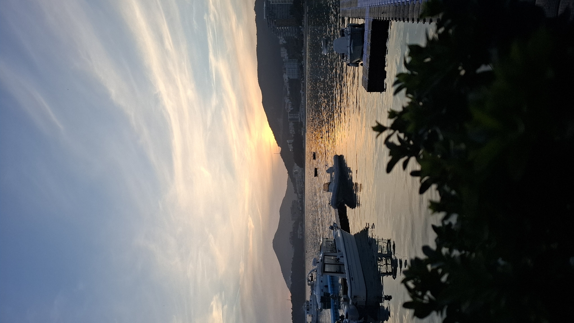 The image depicts a sunset over the Adriatic Sea, likely taken in or near Rafailovići, Montenegro. The sun is setting behind a mountain range, casting a golden glow on the water. Several boats are docked or moored in the foreground and midground, with a pier extending into the water. The sky is a mix of blue and orange hues, with wispy clouds. Foliage is visible in the extreme foreground, partially obscuring the view. The overall scene conveys a tranquil and scenic coastal atmosphere.