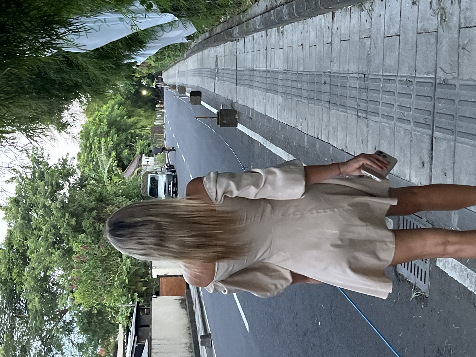 A woman with long blonde hair, dressed in a light-colored dress, walks along a street in the city of Unknown, Indonesia. She holds a mobile phone in her hand. The street is bordered by sidewalks and a row of trees. Cars and other vehicles can be seen in the background. Rectangular wooden blocks are placed along the road.
