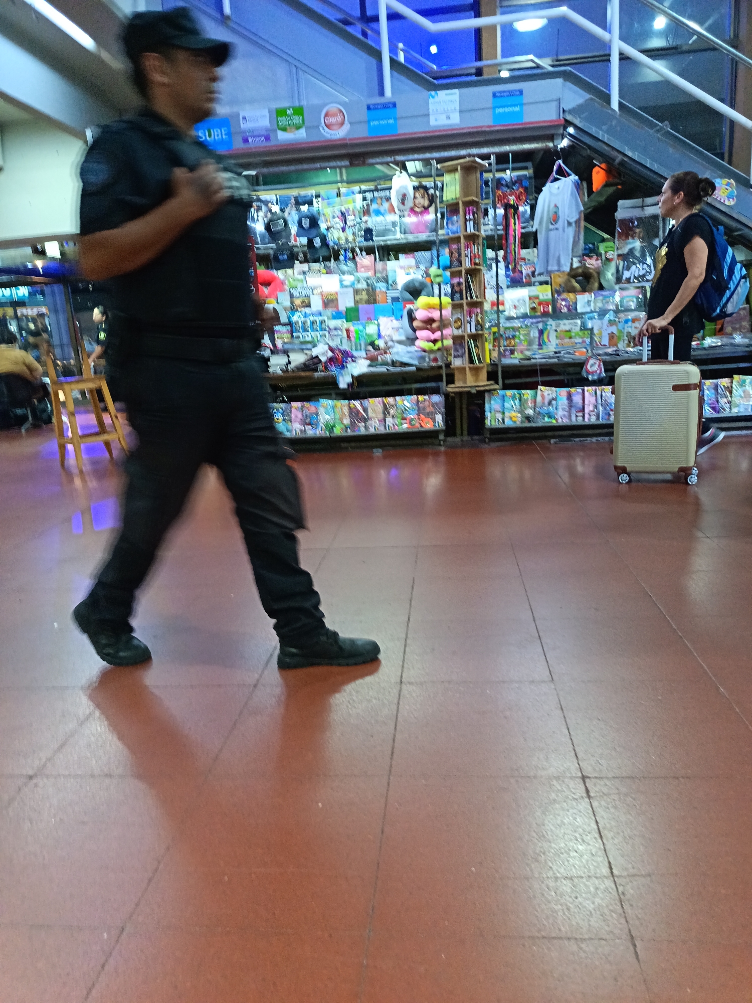 A male security guard walks left across a red-tiled floor in what appears to be the Retiro train station in Buenos Aires, Argentina, given the shops and the signs with the word "SUBE." The guard is wearing a black uniform and a dark cap, and he's in motion. Behind him and to the right, a woman with a blue backpack is standing next to a light-colored wheeled suitcase. Behind them, a store or kiosk is filled with merchandise, including toys, magazines, and souvenirs. An escalator is visible in the background. The lighting is bright and artificial.