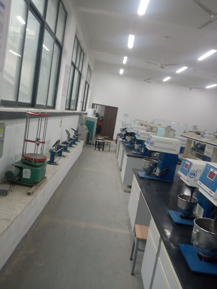 A laboratory room with rows of equipment. On the left, a concrete platform holds testing machines near large windows. The right side features countertops with multiple blue and white mixers. In the background, a doorway leads to another room, with chairs and more equipment visible further down the hall. The lab is brightly lit with overhead fluorescent lights and appears spacious.