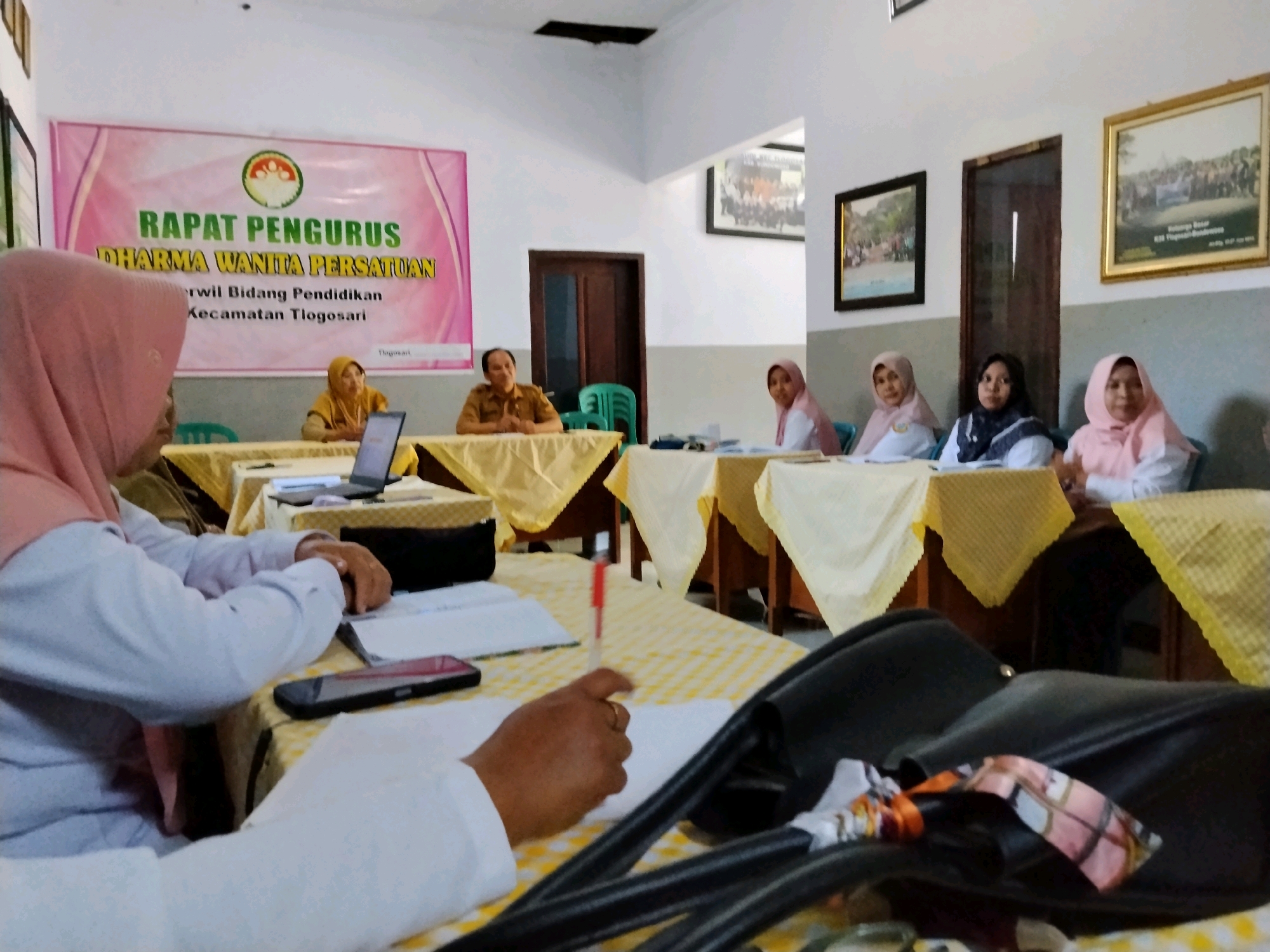 The image captures an indoor meeting, likely a conference room, with a banner in the background that reads "RAPAT PENGURUS DHARMA WANITA PERSATUAN" and mentions "Kecamatan Tlogasari," indicating the location is Tlogasari District. Several people are seated at tables covered with yellow and white checkered tablecloths. The individuals are predominantly women wearing hijabs, suggesting a religious or cultural event. Some are actively participating in the meeting, with papers and pens visible on the tables. In the foreground, a woman in a pink hijab is seen from the back, engaged with documents and a mobile phone. Another person's hand holding a red pen is also prominent. The room appears to be sparsely decorated, with framed pictures on the walls and chairs. The overall atmosphere suggests a formal gathering related to the Dharma Wanita Persatuan, an organization for wives of Indonesian civil servants.
