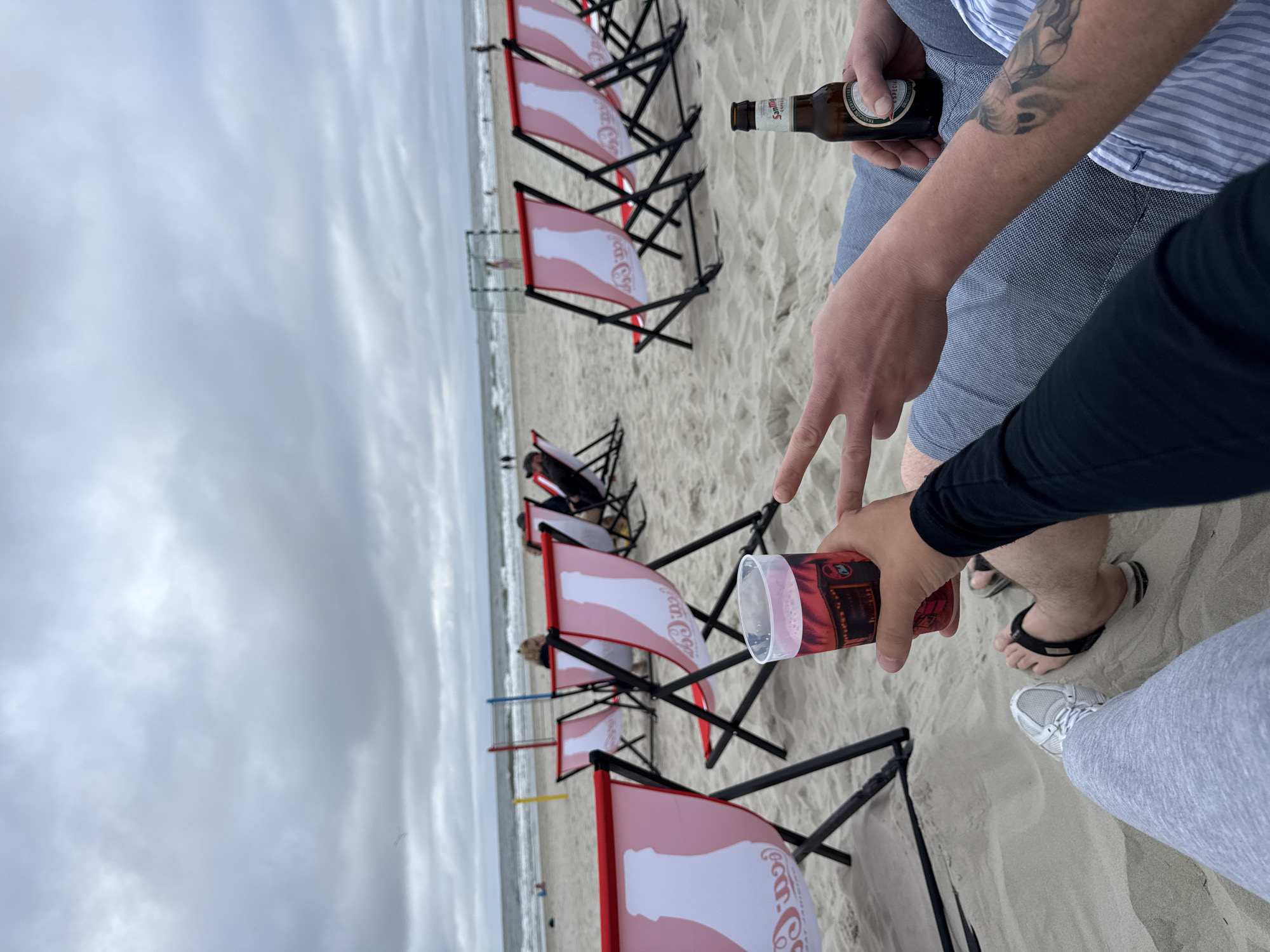 Two individuals are seated on a sandy beach on a cloudy day. In the foreground, one person is holding a clear plastic cup filled with a red beverage and making a peace sign with their hand. Their legs are extended forward, one wearing a grey sneaker and the other a flip-flop. The other person's arm is visible, holding a brown bottle of San Miguel Especial beer. The beach features several red and white striped deck chairs with a Coca-Cola logo. In the background, the ocean is visible, with waves breaking on the shore, and a soccer goal is set up on the sand. The sky is overcast with dense grey clouds. This scene is likely located on the beach in the town of Langeoog, Germany, a North Sea island known for its beaches and recreational activities.