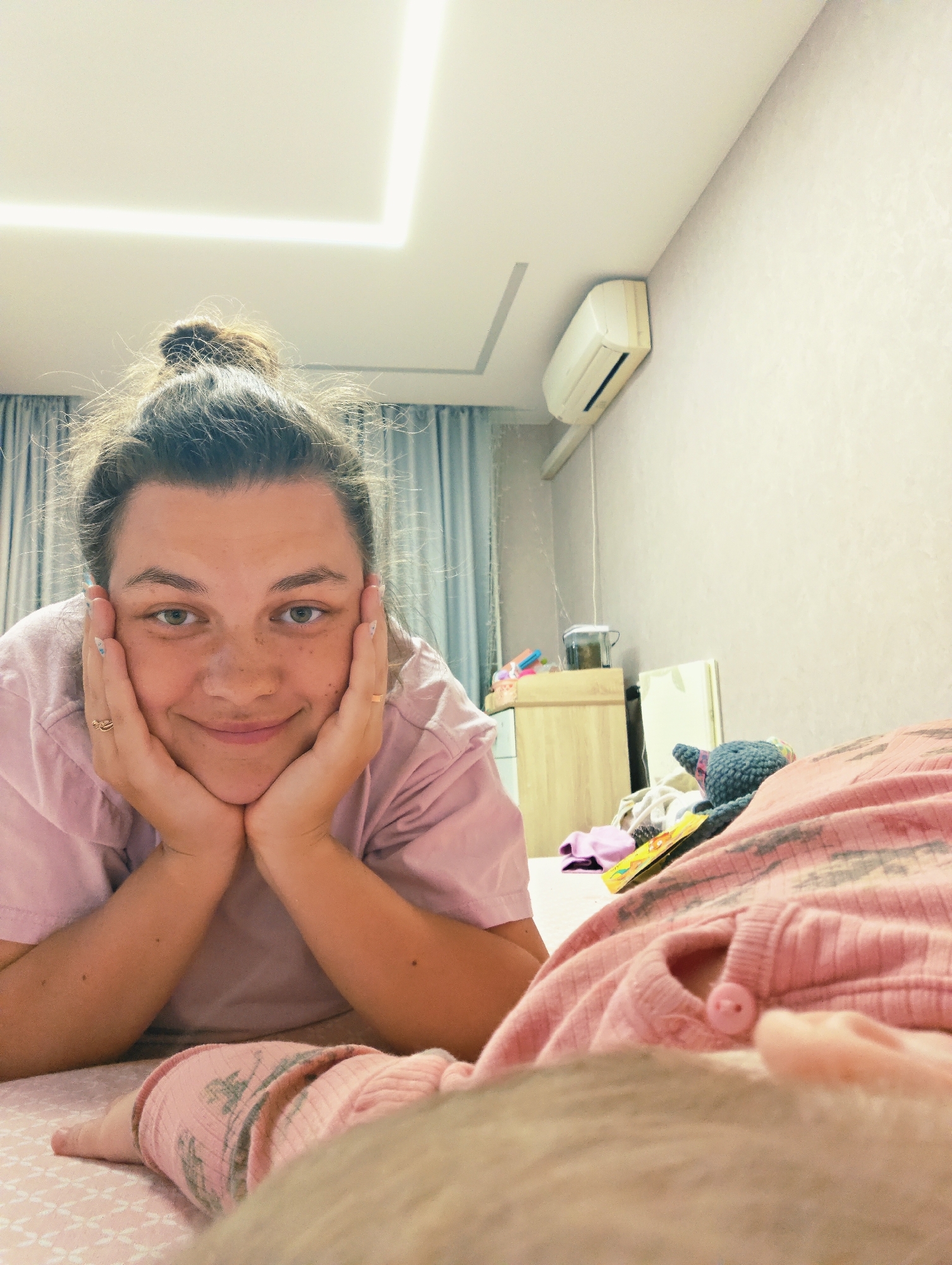 The image depicts a domestic scene in what appears to be a bedroom, located in the city of Chaikovsky, Russia.
A young adult female is the primary subject, positioned in the middle ground, lying prone with her elbows supporting her upper body. Her face is framed by her hands, which rest beneath her chin, exhibiting a gentle smile directed towards the viewer. She has dark hair styled in a high, somewhat dishevelled bun, and her facial features include green eyes and visible freckles across her nose and cheeks. She wears a light pink short-sleeved t-shirt. Two rings are visible on her ring fingers, one on each hand, and her fingernails display light-colored polish with subtle patterns.
In the immediate foreground, largely out of focus and partially obscured, is a small individual, likely an infant or young child, lying on their back. This individual is dressed in pink, ribbed full-body clothing patterned with what appear to be trees or animal silhouettes, and a single pink button is visible on their garment.
The room's environment features light-colored, possibly textured wallpaper on the visible walls. The ceiling is white and incorporates a modern recessed rectangular light fixture. A white, wall-mounted split-unit air conditioner is affixed to the upper right wall. To the left, light blue or grey curtains are partially visible, suggesting a window beyond. A light-colored wooden bedside table or set of drawers stands in the mid-background on the left, topped with various small objects, including a clear plastic container. Further objects, including a dark blue knitted plush toy, some folded fabrics or clothes, and a yellow-covered book, are scattered on the bed or floor to the right of the woman. The overall lighting is warm, emanating from unseen sources and the ceiling fixture.