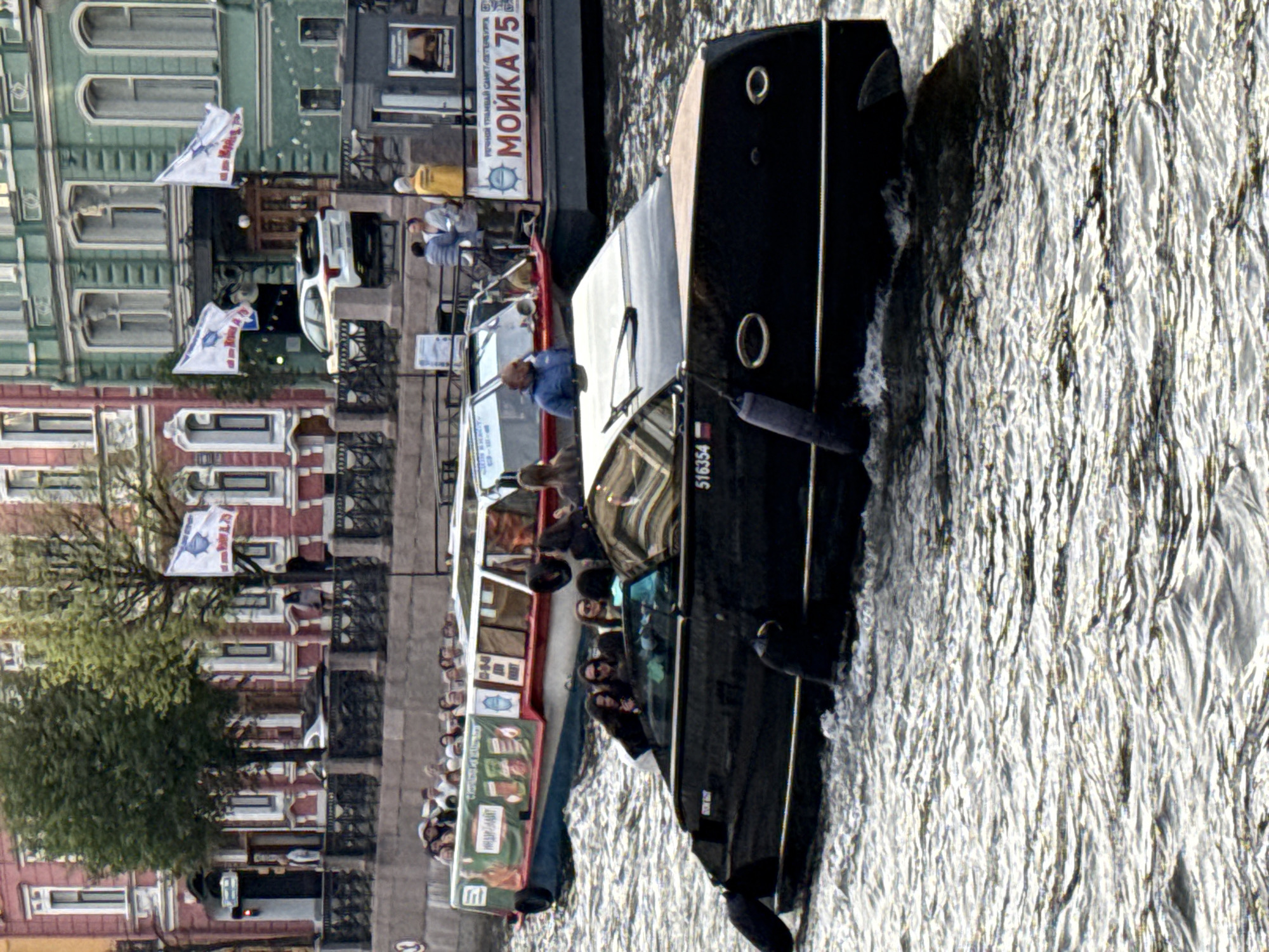 The image depicts a boat tour on a canal in Saint Petersburg, Russia. In the foreground, a black speed boat with several passengers, some appearing to be tourists, navigates the rippling water. The boat has a registration number "516354" visible. In the background, another tour boat, longer and more crowded with passengers, travels in the same direction. Behind the boats, stately buildings with red and green facades line the canal, adorned with white trim and arched windows. Flags are visible flying from some of the buildings. The overall atmosphere suggests a typical day for canal tours in the city.