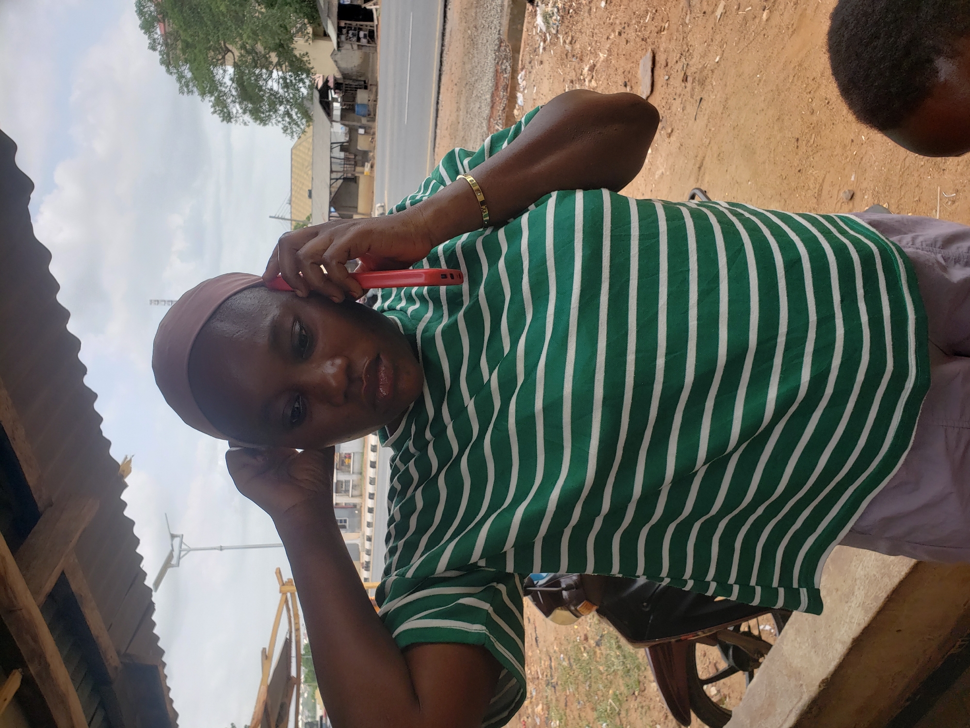The image shows an individual wearing a green and white striped shirt and a head covering, holding a red cell phone to their ear. The setting appears to be outdoors in Adankolo, Nigeria, as indicated by the road and buildings in the background. The individual is wearing a gold bracelet. A portion of another person's head is visible in the upper right corner. The surroundings include a partially visible structure with a corrugated metal roof and a dusty ground surface. A motorcycle is visible in the lower right.