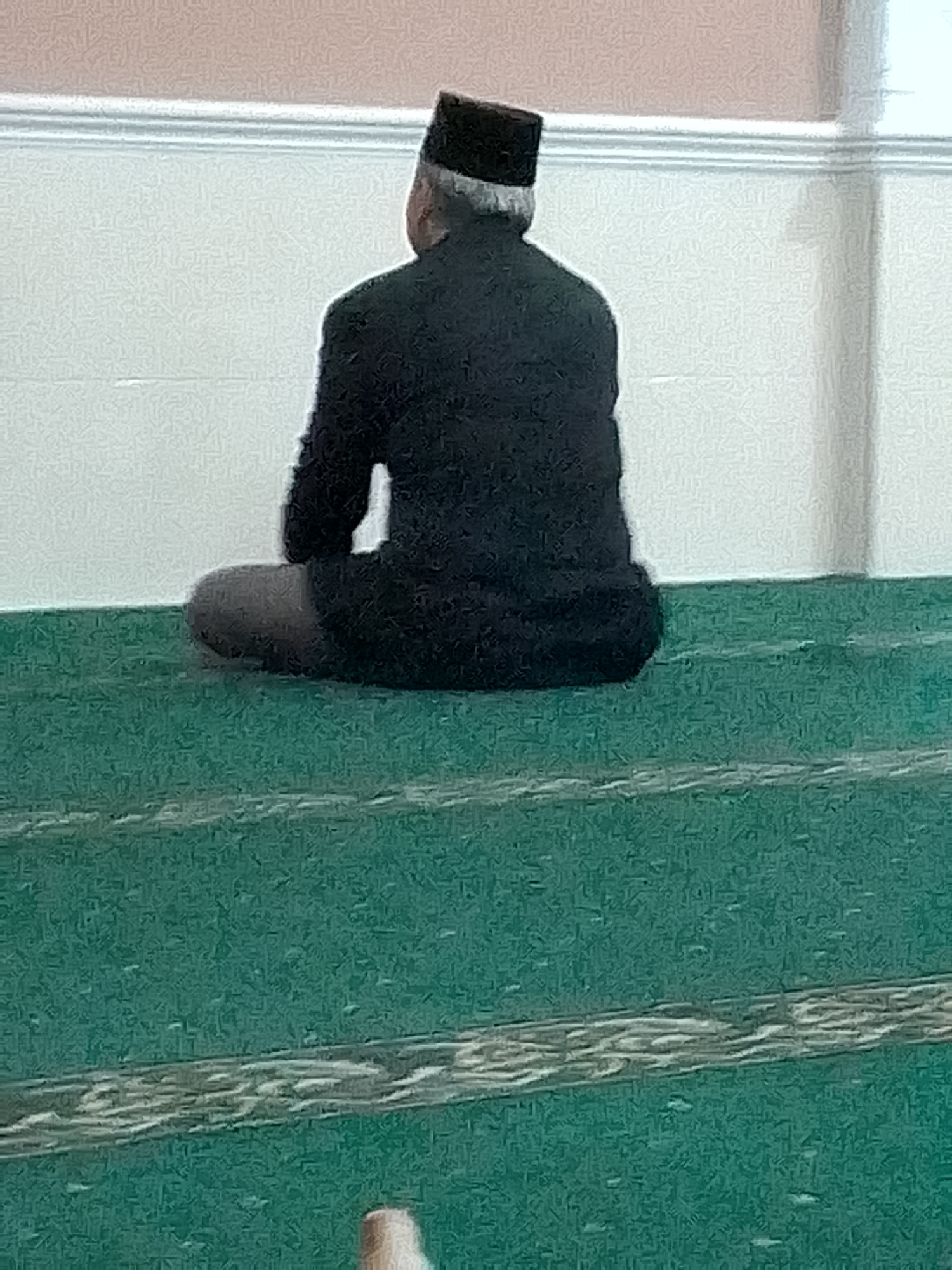 A man in a black robe and a black kufi sits cross-legged on a green carpet with white lines. The carpet is in a room with white walls. The man's back is to the camera, and he is facing a white wall. The man's hair is gray. The man's left leg is visible and appears to be slightly bent. The image was taken in the city of Kertajaya, Indonesia.