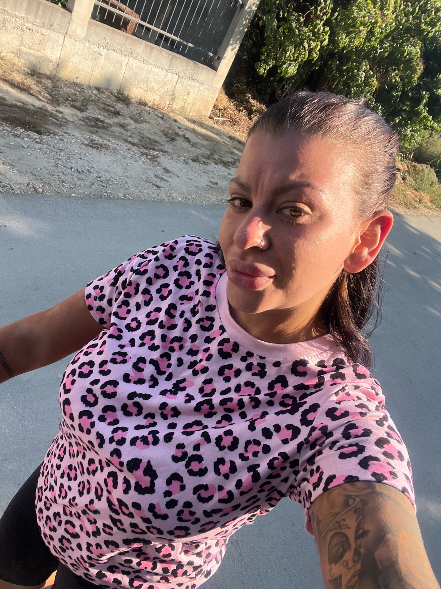 A woman with dark hair, wearing a pink leopard print shirt and black leggings, takes a selfie outdoors. The background features a paved road, a dirt embankment, a concrete wall with a metal fence, and lush green trees under a bright, sunny sky. Her left arm, visible in the foreground, displays tattoos. The scene appears to be in a residential or semi-rural area.