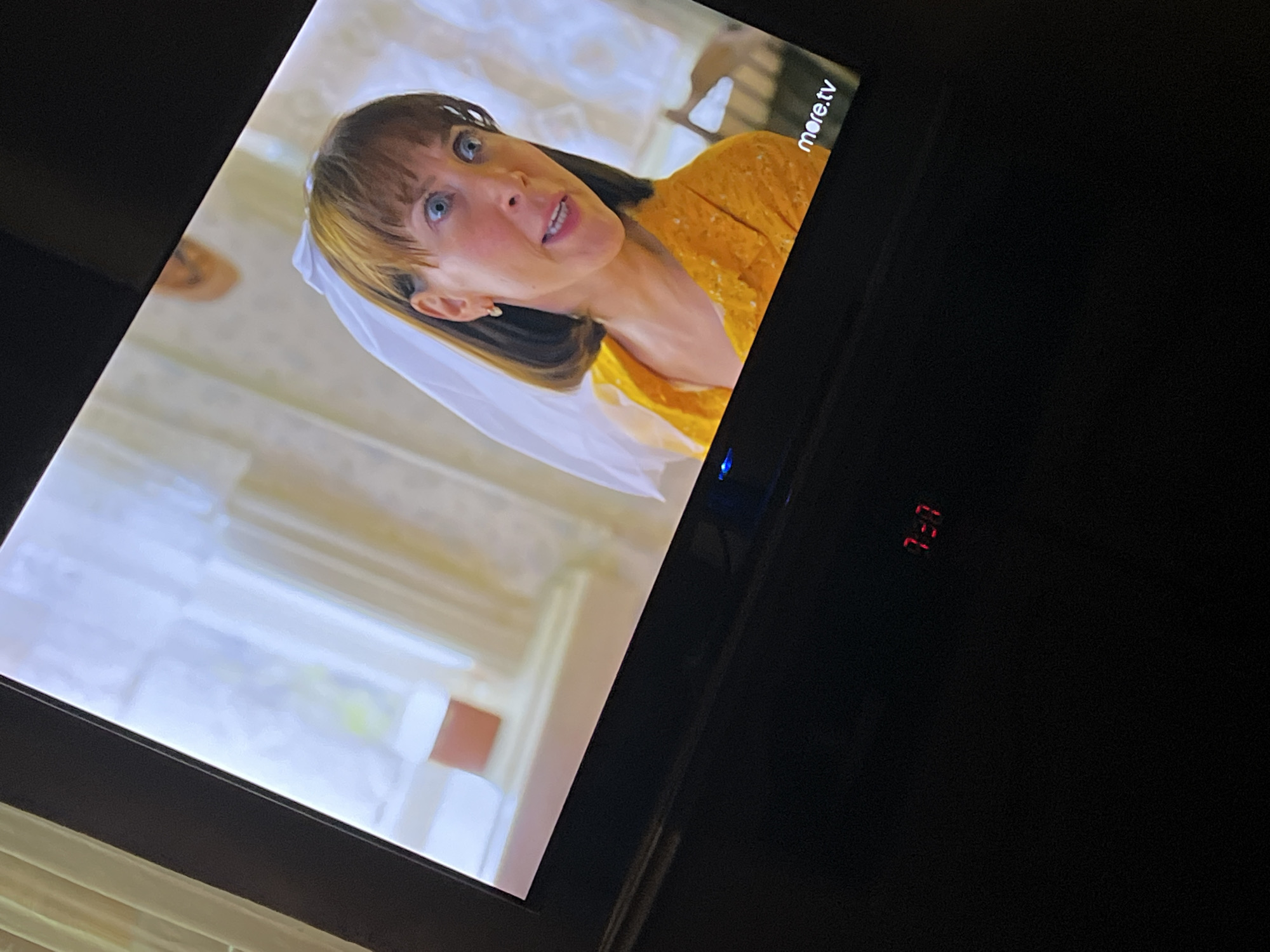 The image shows a television screen displaying a scene with a woman wearing a yellow shirt and a white veil. The background appears to be an interior setting with curtains and furniture. In the lower right corner of the screen, the text "more.tv" is visible. Below the screen, a red digital clock displays "7:07". The image is taken from an angle, suggesting it's a photograph of a television in a dimly lit room. There is no information in the image to suggest a location in Babruysk, Belarus.