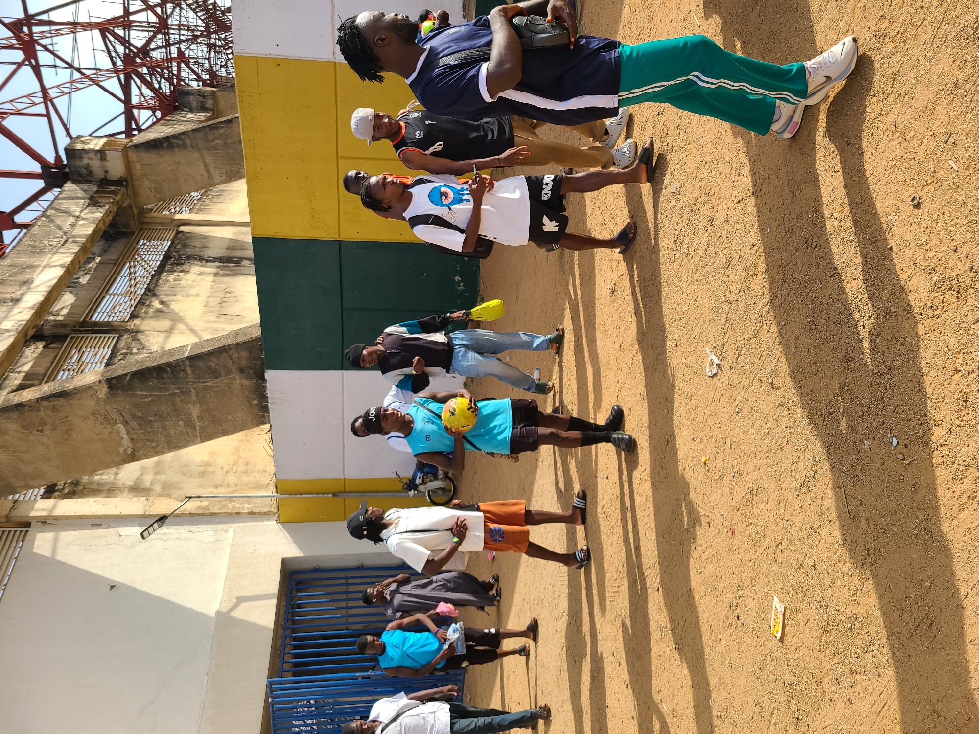 A group of people is standing outside the New Stadium in Nigeria. The stadium structure is visible in the background, including parts of a red metal structure and a concrete wall with rectangular openings. The people are of various ages and are dressed in casual clothing. Some are carrying items such as a soccer ball and bags. The ground is a sandy color with shadows cast by the people and the stadium structure. The scene appears to be outdoors during the daytime.