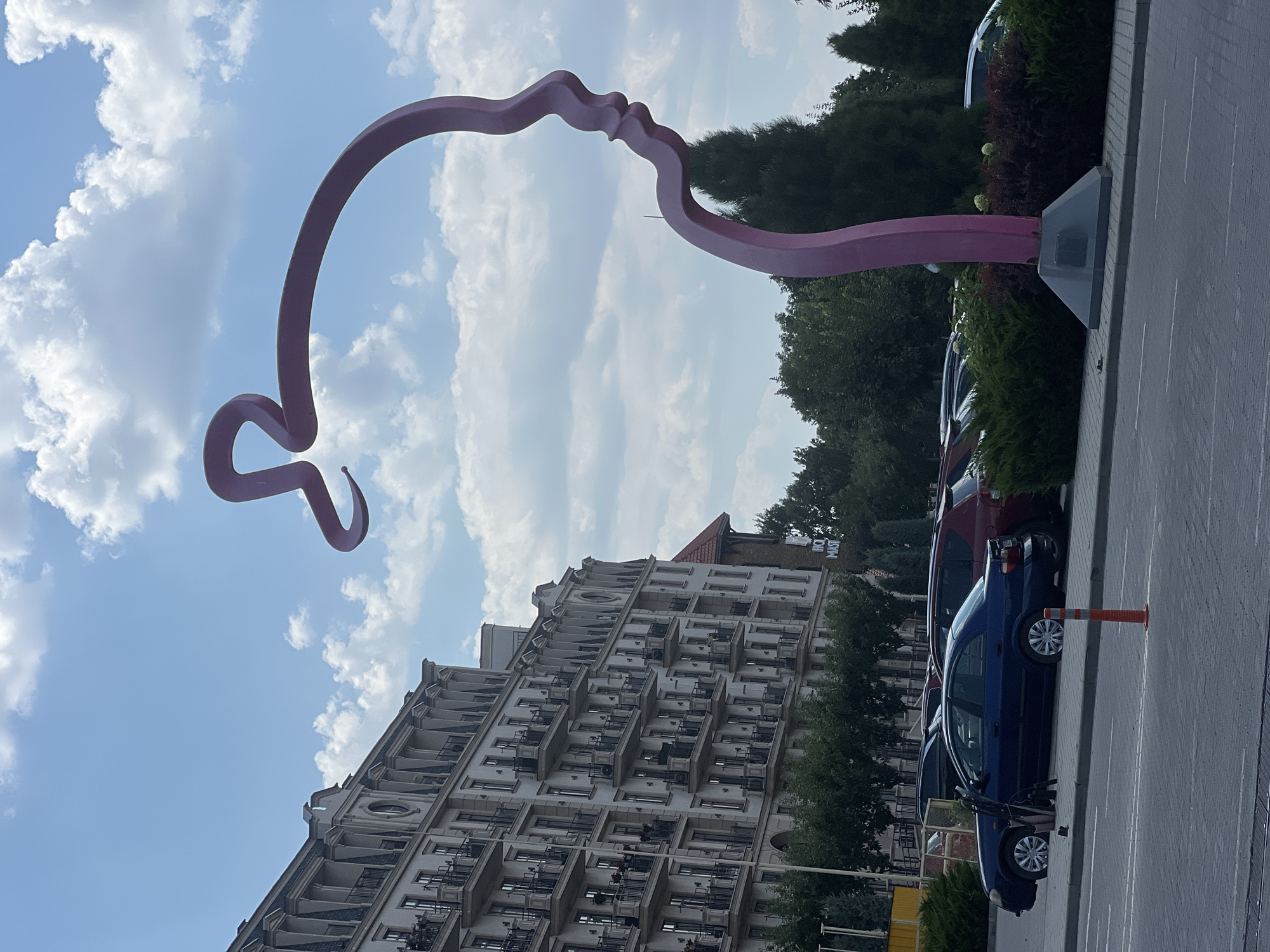 This is a vertical, eye-level shot of a modern outdoor sculpture that resembles a profile of two faces nearly touching. The sculpture is a continuous, smooth, pinkish-red line with a sleek, abstract design. It is set against a bright blue sky with scattered white, puffy clouds, suggesting a pleasant, daytime weather.
In the background, to the left of the sculpture, is a large, multi-story building with a light-colored facade and numerous balconies. The building has a classical architectural style with ornate detailing around the windows and roofline. To the right of the sculpture and the building, there are lush green trees and manicured bushes, indicative of a well-maintained outdoor space.
In the foreground, to the right, a paved surface, likely a parking lot or sidewalk, is visible with white lines marking parking spaces. Several cars are parked along the curb, including a blue hatchback and a darker red SUV. An orange traffic cone stands near the blue car. The scene appears to be in an urban or suburban environment.
The lighting suggests it is daytime, likely mid-morning or afternoon. There is no text clearly visible in the image, except for what might be on the sign in the far background behind the trees, which is too blurry to read. The overall mood is serene and perhaps contemplative due to the sculpture's design. The location context provided is Fontanka, Ukraine.
