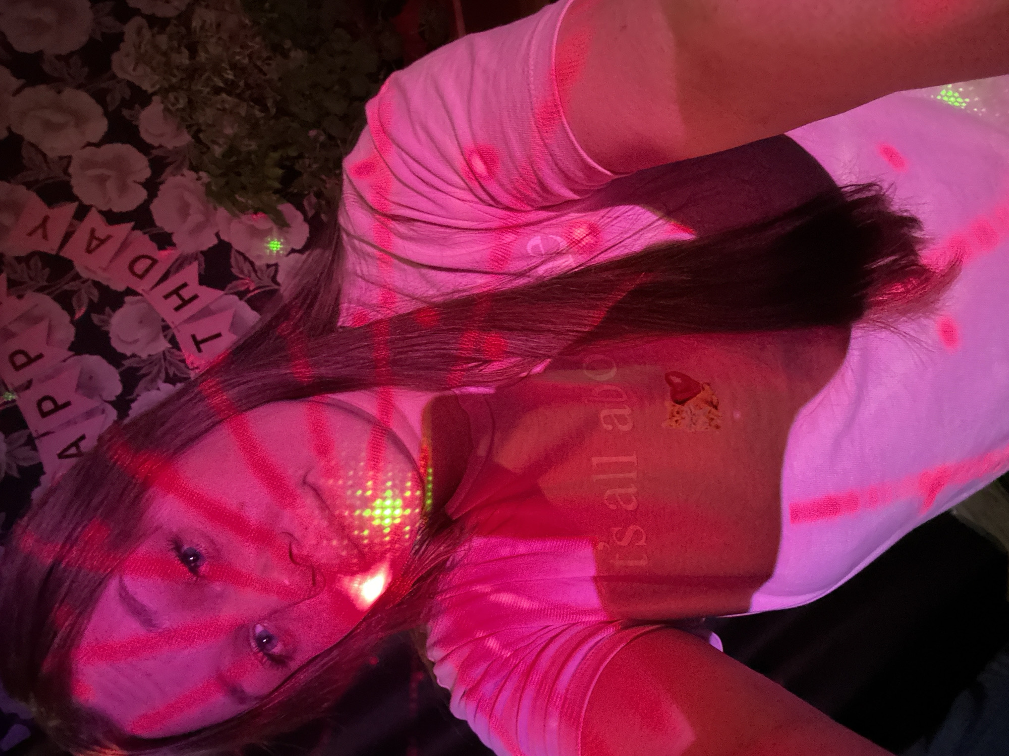 An individual is depicted in a close-up, dimly lit environment in Ольховка, Russia, characterized by ambient red and pink light and projected green laser dots. The subject is a female, likely a young adult, with long dark hair, wearing a light-colored (appearing pink due to lighting) short-sleeved t-shirt. The shirt features text partially visible as "it's all abou" and a small graphic, possibly a cat with a heart, on the upper chest. Her face is illuminated with strong red and pink light, and green laser dots are visible across her face, particularly on her nose and chin, and on her upper body. She is looking directly towards the camera. The background reveals a dark wall with a white floral pattern and a celebratory banner spelling "HAPPY BIRTHDAY" (partially visible as "HAP" and "THDAY"). A dark green plant or foliage is also visible in the background, near the banner.