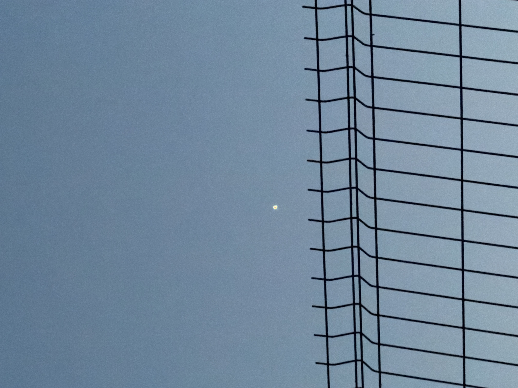 A nighttime outdoor photograph captures a clear, pale blue sky above a black metal fence. The fence, made of wire mesh with a top horizontal rail and vertical wires, occupies the lower half of the frame. A single bright, yellow-white light source is visible in the sky, approximately in the center and slightly above the fence line. The sky is otherwise devoid of clouds or other celestial bodies. The scene appears to be a public space, possibly a park or an industrial area, given the presence of the fence. No people or animals are present in the image.