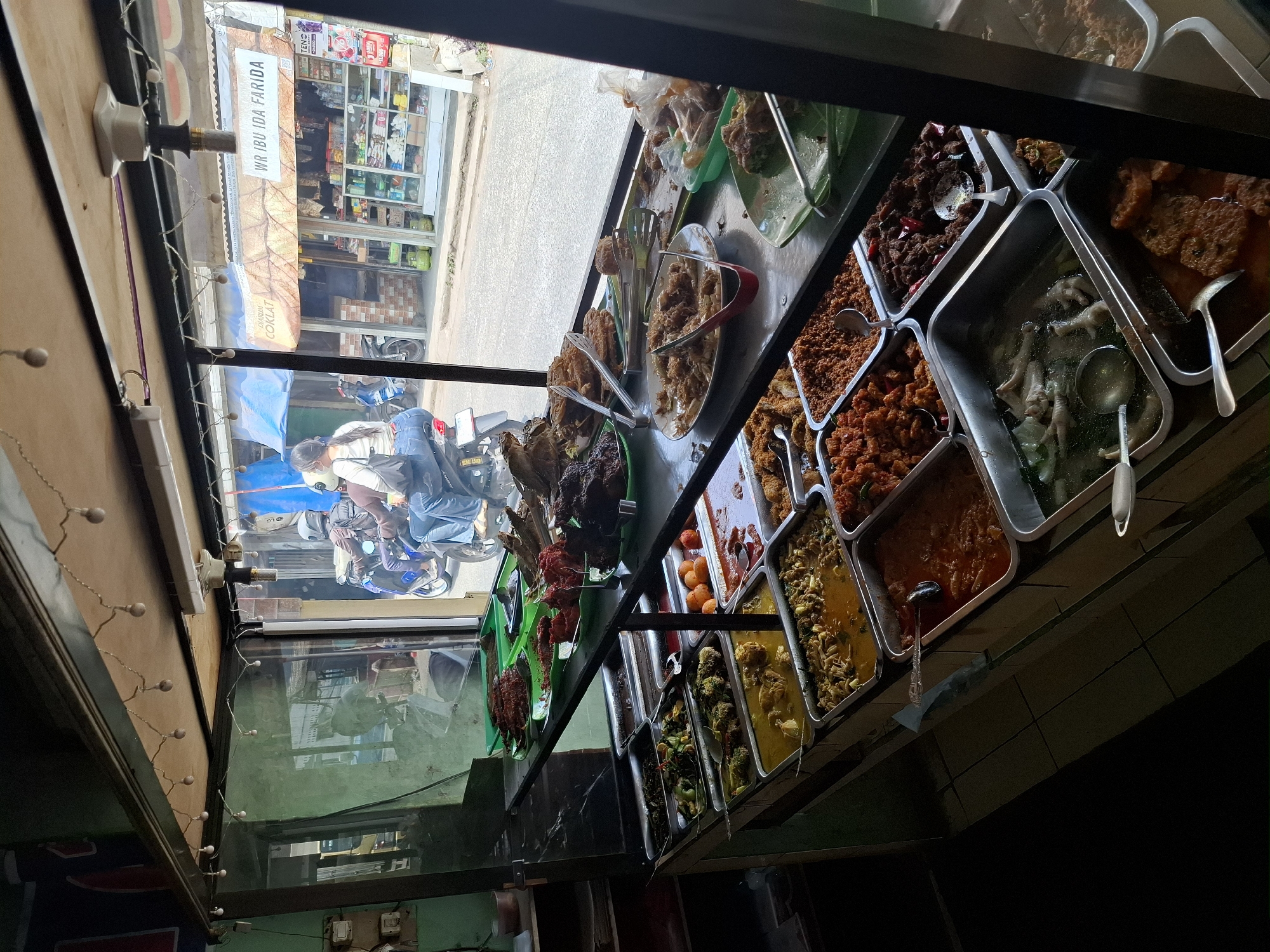 This is a medium-shot, eye-level, natural light photograph of a food stall in Cibadak, Indonesia, seen from the interior of the stall looking out. The stall displays a variety of Indonesian dishes in rectangular metal trays arranged on shelves. The food includes curries, stir-fried vegetables, fried items, and what appear to be chicken feet in broth. Tongs are placed in several of the trays for serving.
In the background, visible through a glass window, is a street scene. A woman wearing blue jeans and a white shirt is riding on the back of a blue motorcycle, which is driven by a person wearing a helmet. The motorcycle is stopped, with other motorcycles and people visible on the street. A shop with signage that reads "WR IBU IDA FARIDA" and "COKLAT" (chocolate) is situated across the street, with shelves stocked with various products. The overall environment suggests a busy, local marketplace or street food area. The stall itself has a simple metal frame structure with a wooden top. Strings of small, bare lightbulbs are attached to the underside of the wooden top, providing illumination within the stall.