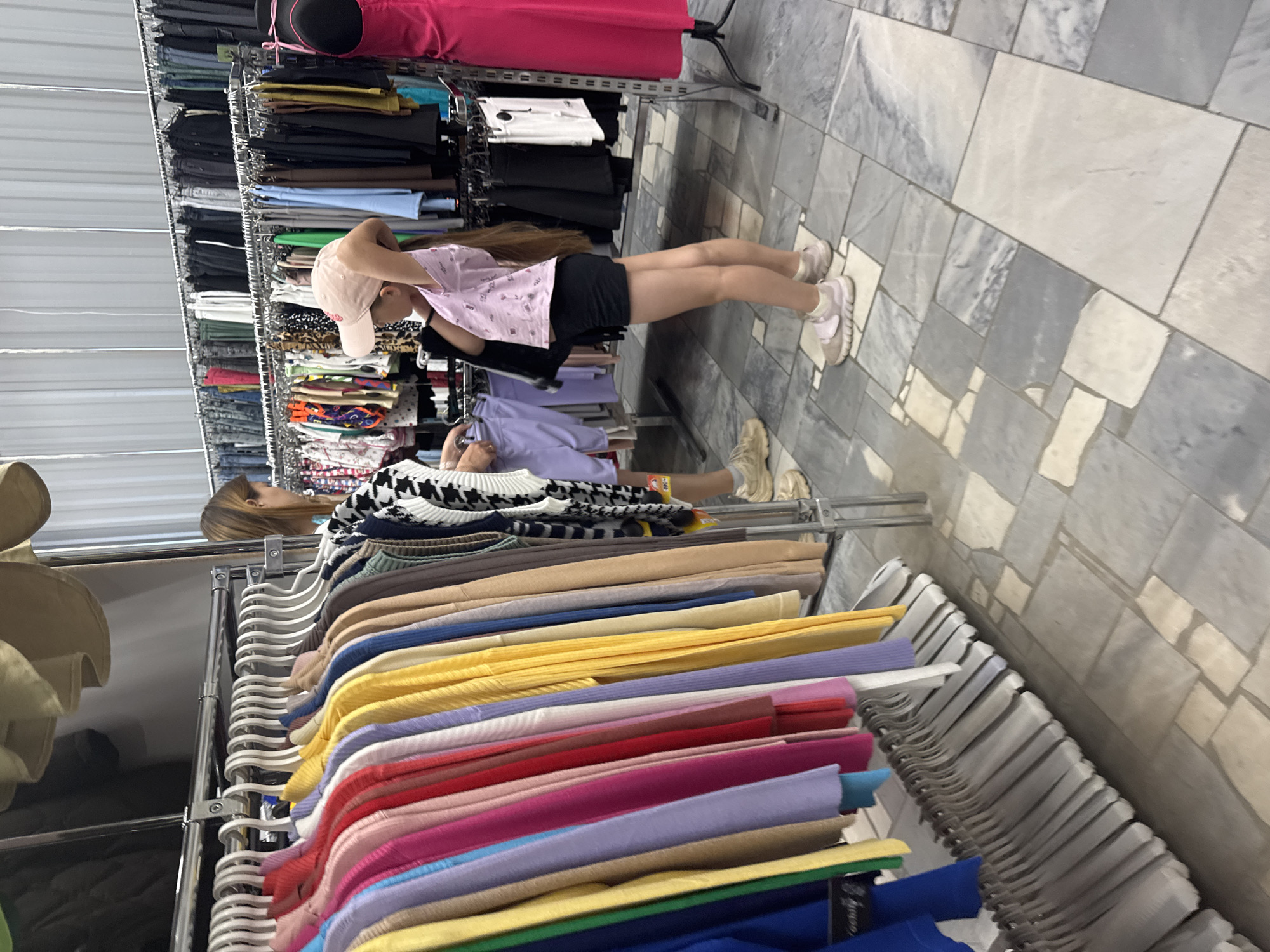 The image depicts the interior of a clothing store. Two people are present. One person, wearing a pink shirt, black shorts, a light pink cap, and pink shoes, is standing and looking at clothing racks. The other person, wearing a black and white sweater and lilac pants, is also looking at the clothing racks. The clothing racks are filled with clothes of various colors, including yellow, pink, and blue. The floor is tiled in a gray and white pattern. The store is located in the city of Chaikovsky, Russia.