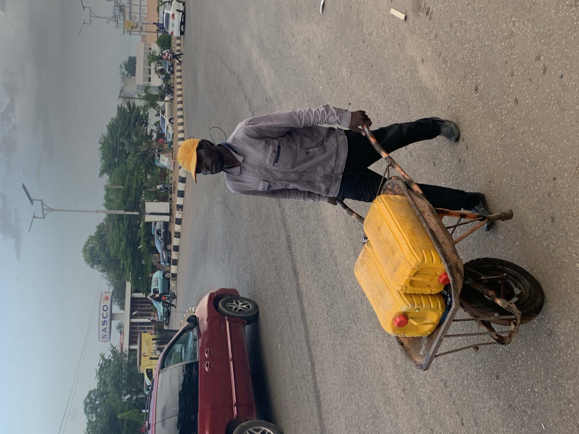 In the city of Jos, Nigeria, a man wearing an orange cap and a grey jacket pushes a wheelbarrow filled with two large yellow jerrycans with red caps. The wheelbarrow has a rusty orange frame and a single black tire. The man is walking on a paved road with a red minivan in the background. The sky is overcast. In the background, there is a sign that reads "NASCO" and other buildings and trees. There are also other vehicles and people present on the street.