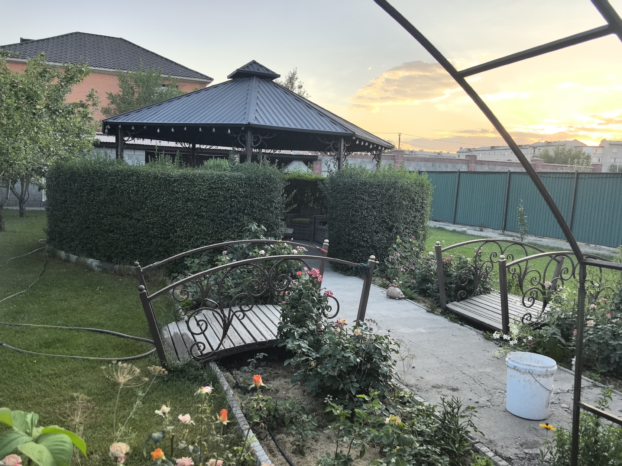 The image depicts a backyard scene at dusk, likely in Taldykorgan, Kazakhstan. In the foreground, a small ornamental bridge with wooden planks and decorative metalwork spans a dry garden bed. Roses and other greenery surround the bridge. To the left, a green lawn is partially covered by a garden hose. Behind the bridge and garden, a neatly trimmed hedge encloses a larger area. Beyond the hedge, a dark-colored, multi-sided gazebo with a pointed roof stands. In the background, a brick building with a dark tiled roof is visible, along with a green metal fence. The sky is overcast with soft, warm light indicating the setting sun. The overall impression is of a well-maintained private garden.