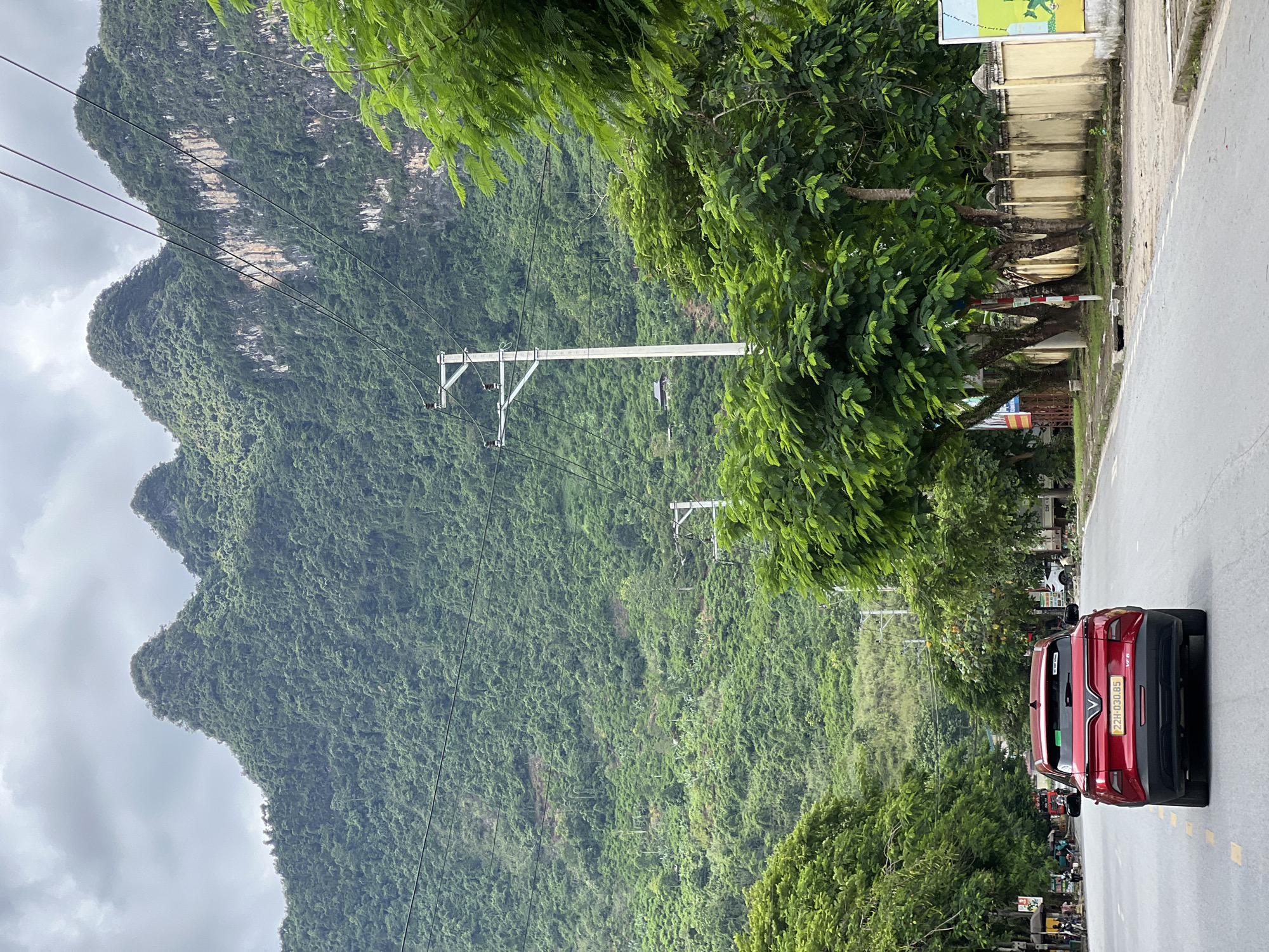 The image shows a road in the city of Hà Giang 2 Ward, Vietnam, with a red car driving away from the camera in the foreground. The road is lined with trees on both sides, and a large, lush green mountain range dominates the background. The sky is overcast. There are power lines and utility poles visible along the road. In the distance, there are some buildings and more vegetation on the mountainside. There are a few people and motorcycles on the side of the road in the far distance, near some market stalls. A yellow wall with decorative elements runs along the right side of the road. The overall scene depicts a scenic road in a mountainous region.