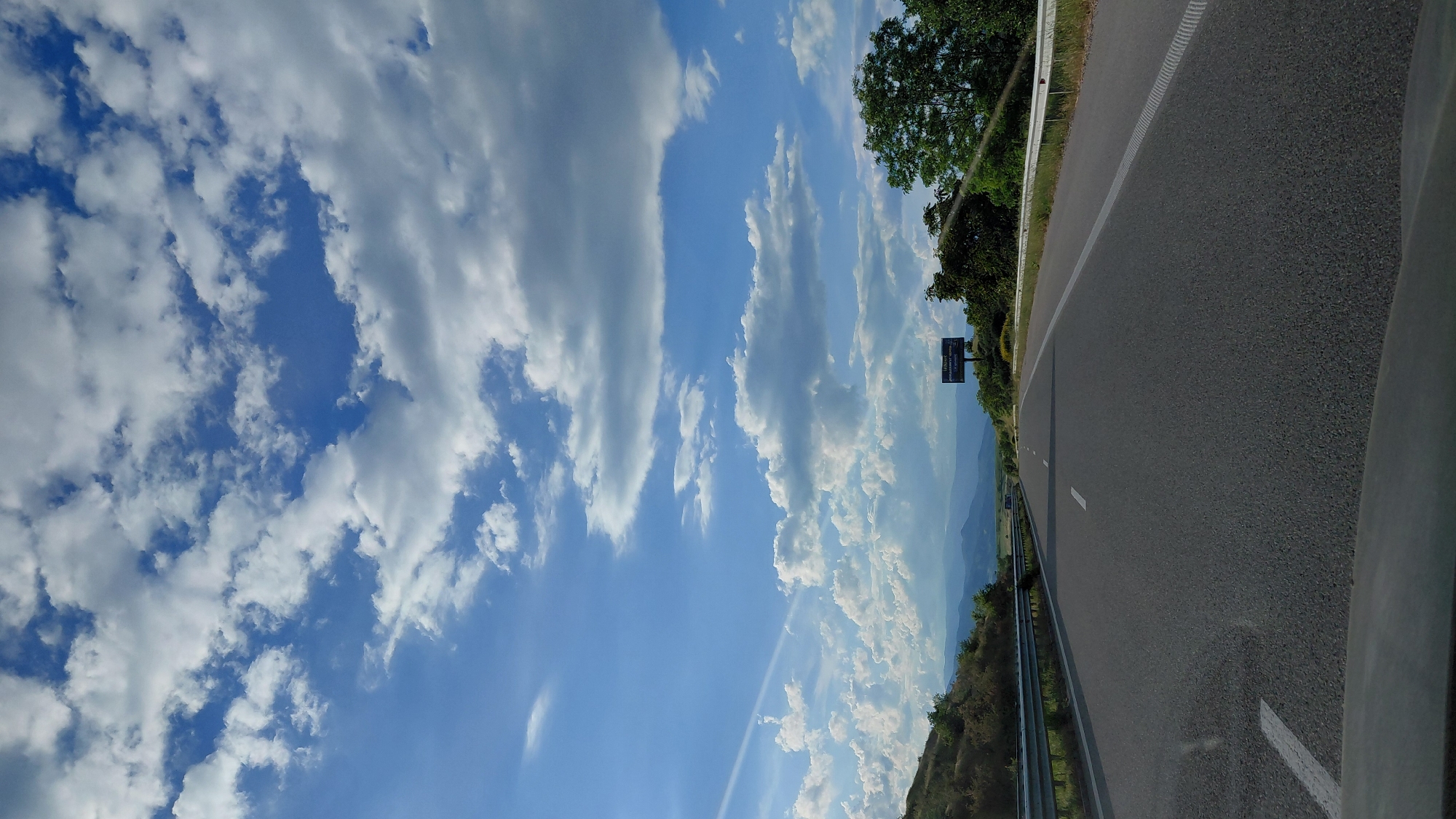 The image shows a view from a vehicle moving along a highway. The sky is predominantly blue with scattered white clouds. To the right, the highway stretches into the distance with a guardrail visible. On the side of the road, there is some vegetation and a highway sign. In the distance, there are hints of mountains. The location is Unknown, Unknown.
