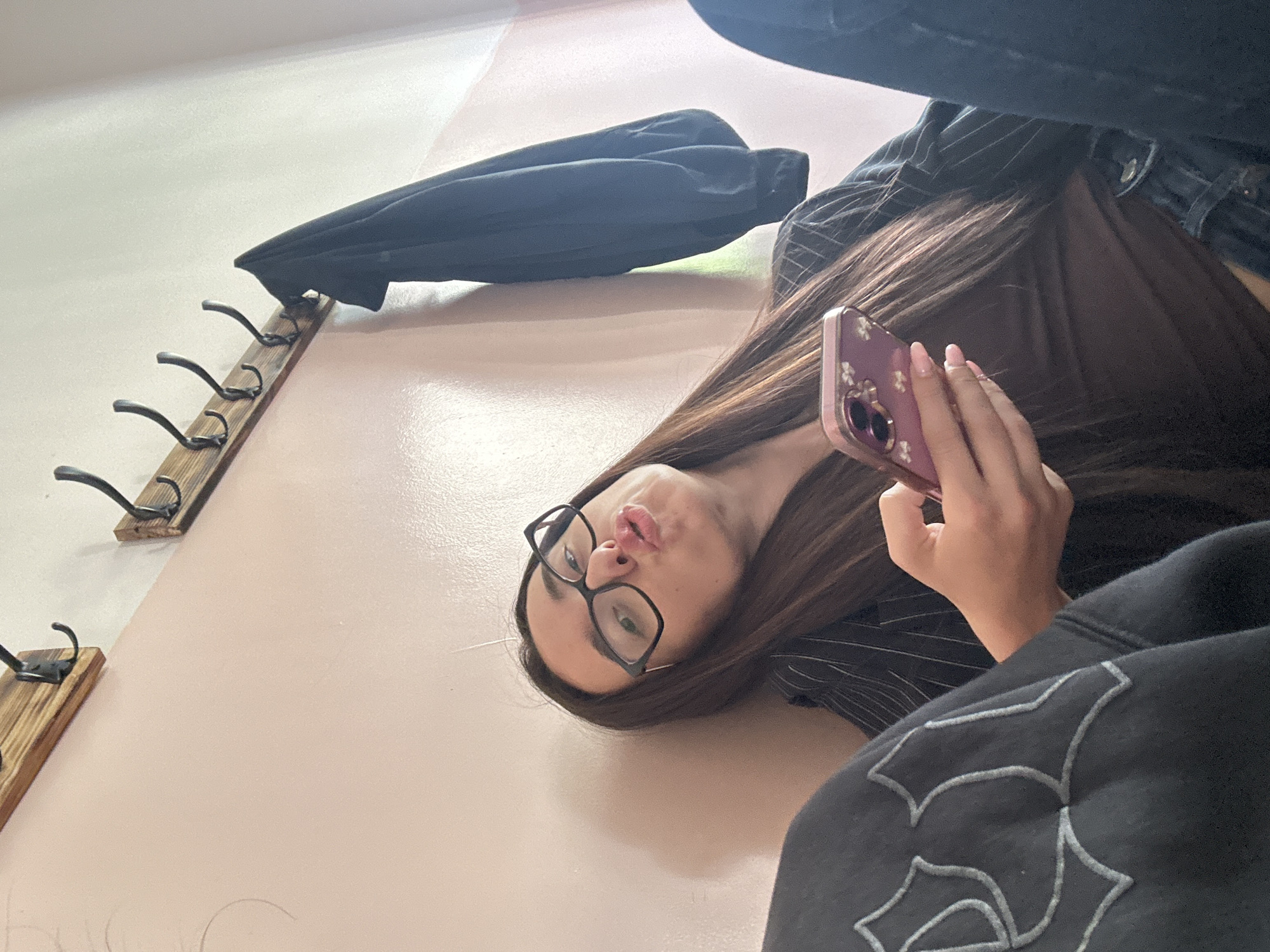 The image captures a casual, indoor scene in Sofia, Bulgaria, featuring a young woman taking a selfie or posing for a photo. The perspective is from below, looking up at the subject, who appears to be lying down or leaning back.
The primary subject is a young woman with long, straight, dark brown hair. She is wearing black-rimmed glasses and her lips are pursed in a playful "duck face" or blowing a kiss expression. She is dressed in a black top with subtle white pinstripes, covered partially by a black hoodie featuring a distinctive grey, possibly gothic or abstract, embroidered or printed design on the sleeve. Her nails are manicured with pink polish. She is holding a pink smartphone, encased in a floral-patterned case, in her right hand, suggesting she is interacting with the device, likely taking the photo.
The setting appears to be a room with walls painted in two tones: a light beige or off-white on the upper section and a light pastel pink below. A wooden coat rack with five dark metal hooks is visible on the wall, extending from the bottom left corner upwards. A black garment, possibly a jacket or coat, is draped over one of the hooks. Another folded black garment, perhaps trousers, is visible lying on the floor or surface near the woman.
The overall mood is lighthearted and personal. There are no specific cues within the image to definitively confirm the location as Sofia, Bulgaria, beyond the provided context. The time of day is not evident, but the lighting suggests an indoor setting, possibly with ambient artificial or natural light. No discernible text is visible on any object.