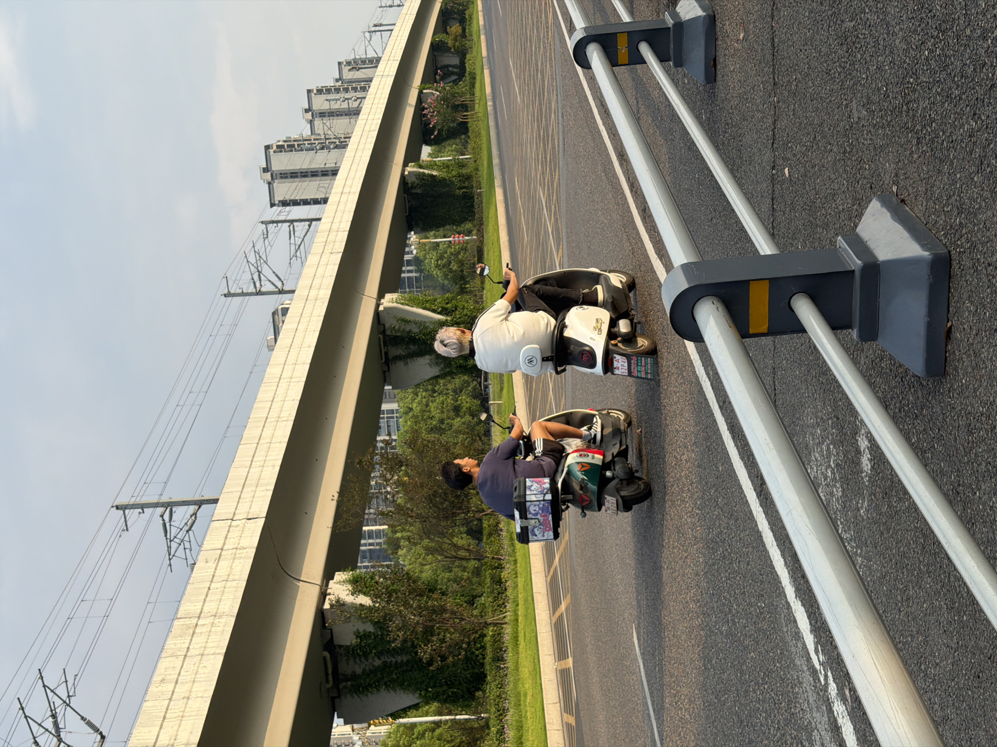 This image captures a street scene in Jiangjin, China, on a clear day. Two individuals are riding electric scooters in the same direction on a paved road. The scooter on the left, a green and white model, is ridden by a person wearing a dark t-shirt and shorts. Attached to the back of this scooter is a white storage box adorned with cartoon characters. The scooter on the right, a white model, is ridden by a person wearing a white t-shirt and black pants, with their back to the camera. Both scooters have license plates visible.
In the background, a prominent elevated structure, likely a viaduct or overpass, spans across the scene. It is supported by concrete pillars, some of which are covered in greenery. Overhead, power lines and their supporting infrastructure are visible. Beyond the overpass, several multi-story buildings, characteristic of urban development, rise against the sky. The road itself features lane markings and a silver metal guardrail with dark gray posts runs along the right side of the image, separating the road from an unseen area. The lighting suggests it is late afternoon or early evening, with a warm glow illuminating the scene.
