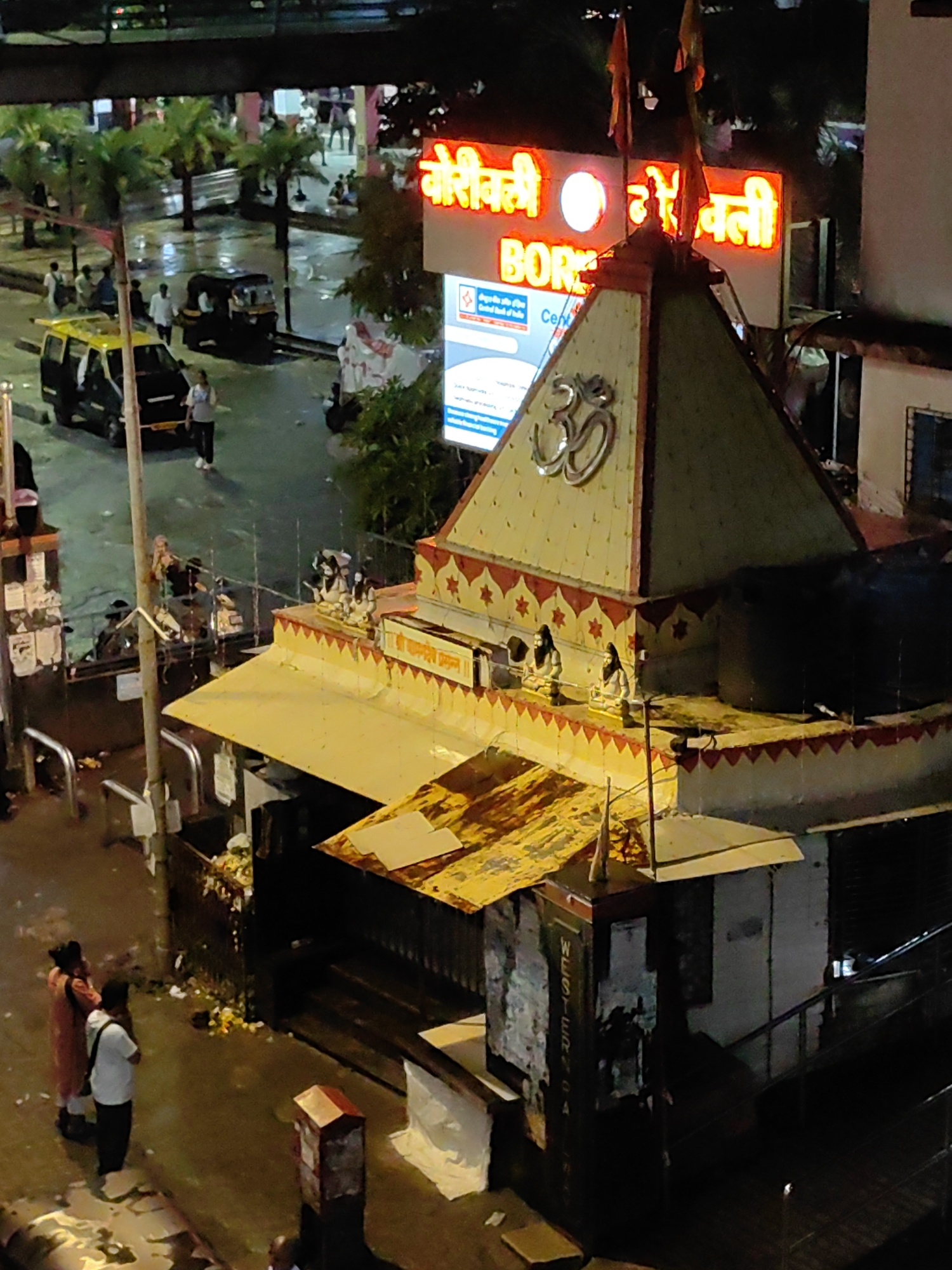 The image captures a street scene in Mumbai, India, at night, likely near a religious site or market, as indicated by the presence of a temple-like structure and illuminated signs. In the foreground, two individuals, one wearing a pink outfit and the other a white shirt and black pants, stand facing away from the camera. A small, red-topped structure, possibly a post box or information kiosk, is visible to their right.
The central focus of the image is a temple structure with tiered roofs and decorative elements. A prominent "Om" symbol is mounted on the peak. Several statues of deities or figures are placed on the temple's ledges. Above the temple, illuminated signs in a reddish-orange neon glow are visible. One sign reads "BOR" and another appears to contain Marathi script. Below these, a blue and white sign, possibly for a bank, is partially visible.
The street is wet, suggesting recent rain, and reflects the surrounding lights. Several vehicles are present, including a yellow and black auto-rickshaw and a van. Palm trees and other greenery are scattered throughout the scene, typical of Mumbai's urban landscape. In the background, more people are visible, some sitting and others standing, and buildings form the backdrop. The overall atmosphere is lively and characteristic of a busy Indian city at night. The sign with "WESTERN" is visible on a dark structure on the right side of the image.
