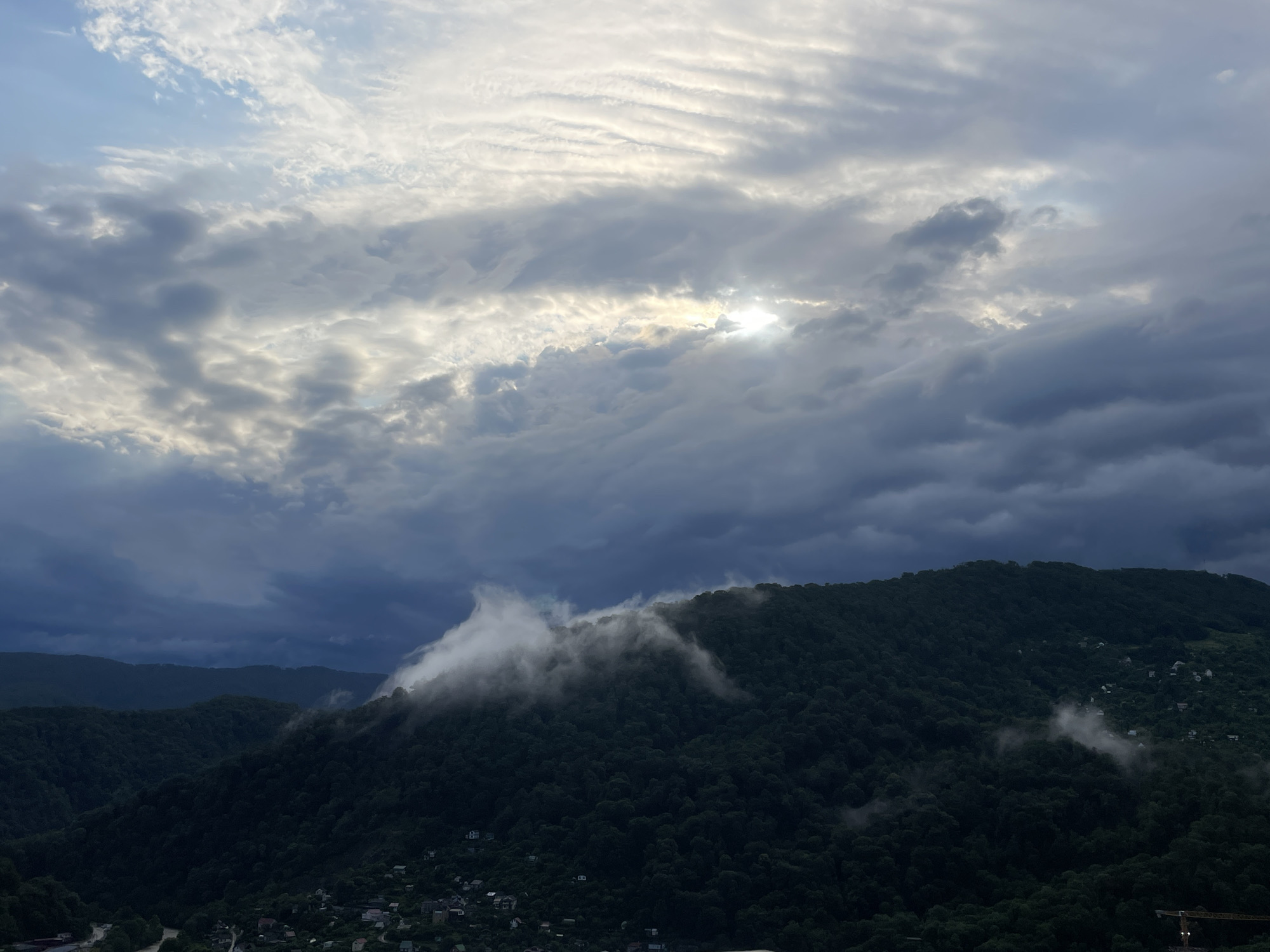 The image depicts a mountainous landscape under a cloudy sky, likely at dusk or dawn given the lighting. The foreground features a dense, dark green forest covering the slopes of the mountains. Small settlements with houses are visible scattered among the trees, particularly on the lower slopes. A winding road or river is faintly visible in the bottom left. In the middle ground, clouds are seen clinging to the mountain peaks, creating a misty and atmospheric effect. The sky is filled with a mix of dark, heavy clouds and lighter, more diffused clouds, with the sun peeking through in the upper center, casting a soft glow. The overall scene conveys a sense of natural beauty and tranquility, with a hint of the dramatic due to the weather. The provided context indicates the location is Baranovka, Russia, which is a resort town in the Krasnodar Krai region known for its mountainous terrain and proximity to the Black Sea.