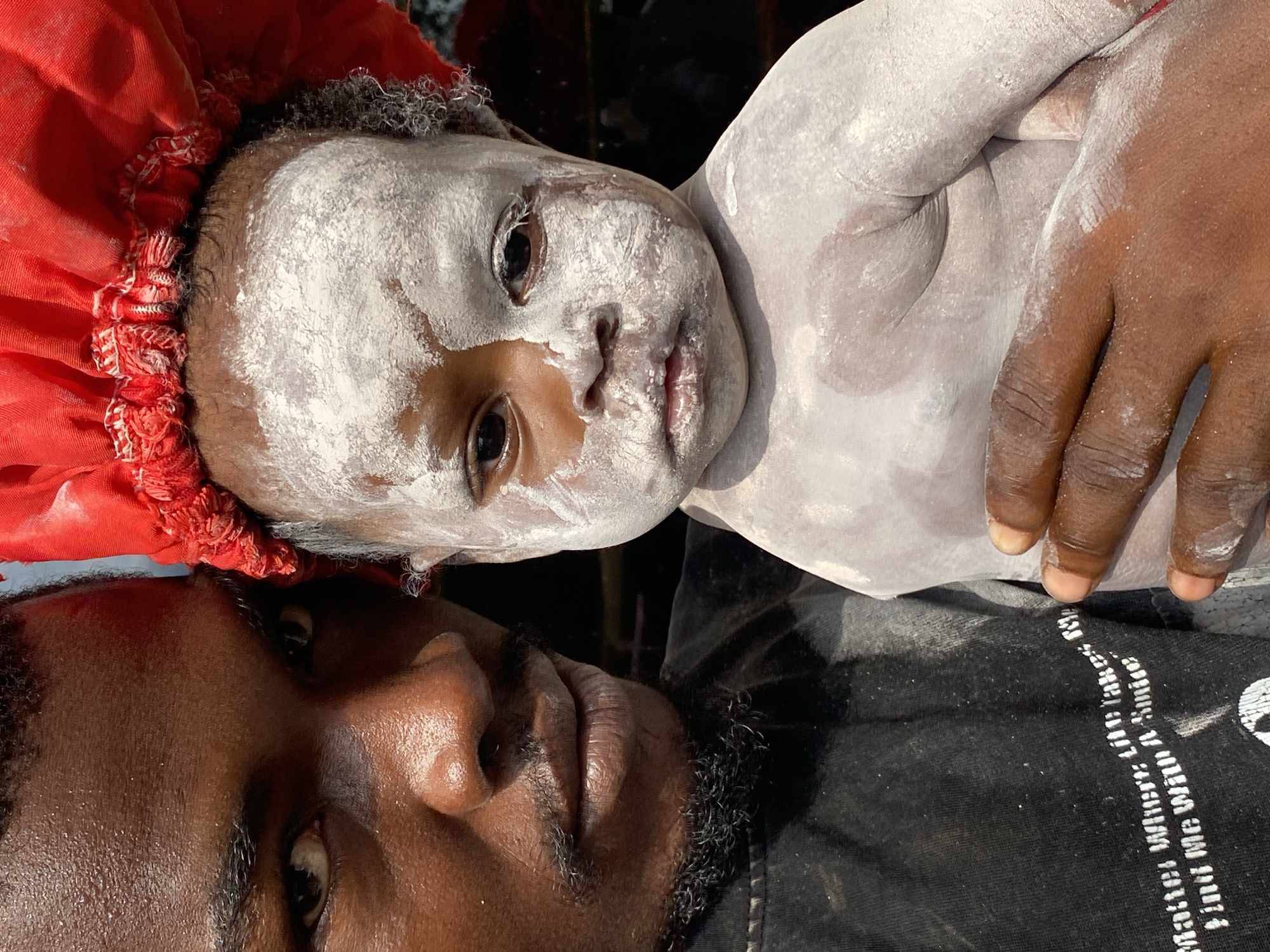 In a close-up, outdoor portrait, a dark-skinned man holds a baby coated in white powder. The man's face is partially visible on the left, with his eye looking directly at the camera, sweat glistening on his forehead. He has a thin mustache and beard. The baby, in the center and right of the frame, is covered in white powder, with some streaks on its face. The baby wears a red cloth head covering. The man is wearing a black t-shirt with white text that reads "Matter Where Life Takes Me, Find Me With A Smile". The background is dark and indistinct, suggesting an outdoor setting. The image appears to have been taken in Ufu Esioni, Nigeria.