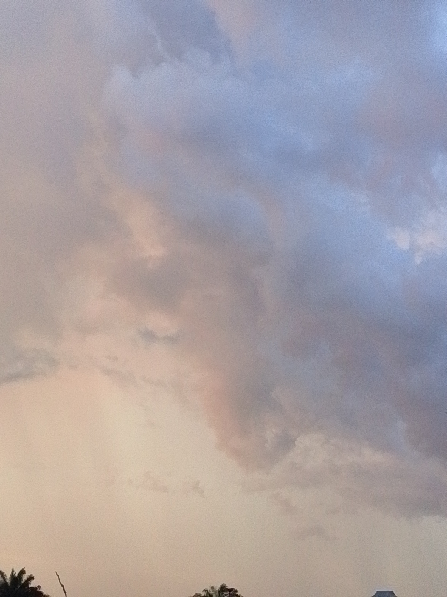 The image shows a cloudy sky with the sun setting in Serti, Nigeria. The clouds are a mix of pink, purple, and blue. The bottom of the image shows the silhouettes of trees and a building.