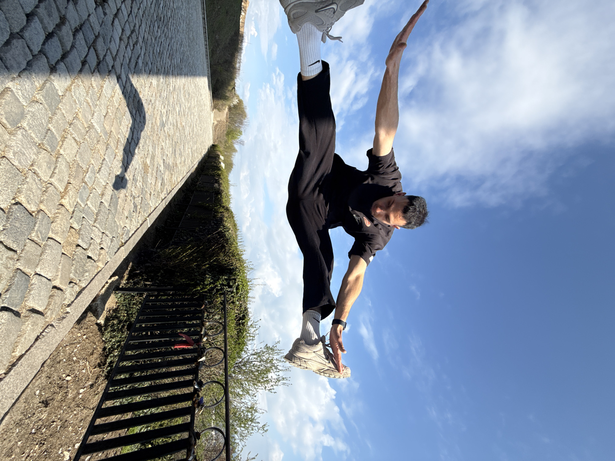A man is performing a gymnastic move outdoors on a sunny day with a blue sky and scattered clouds. He is wearing black pants, a black t-shirt, white socks with the Nike logo, and gray sneakers. He is standing on one leg with his other leg extended upwards and his arms reaching up. The background includes a stone walkway and a black metal fence with shrubbery behind it. This image appears to be taken in Shumen, Bulgaria.